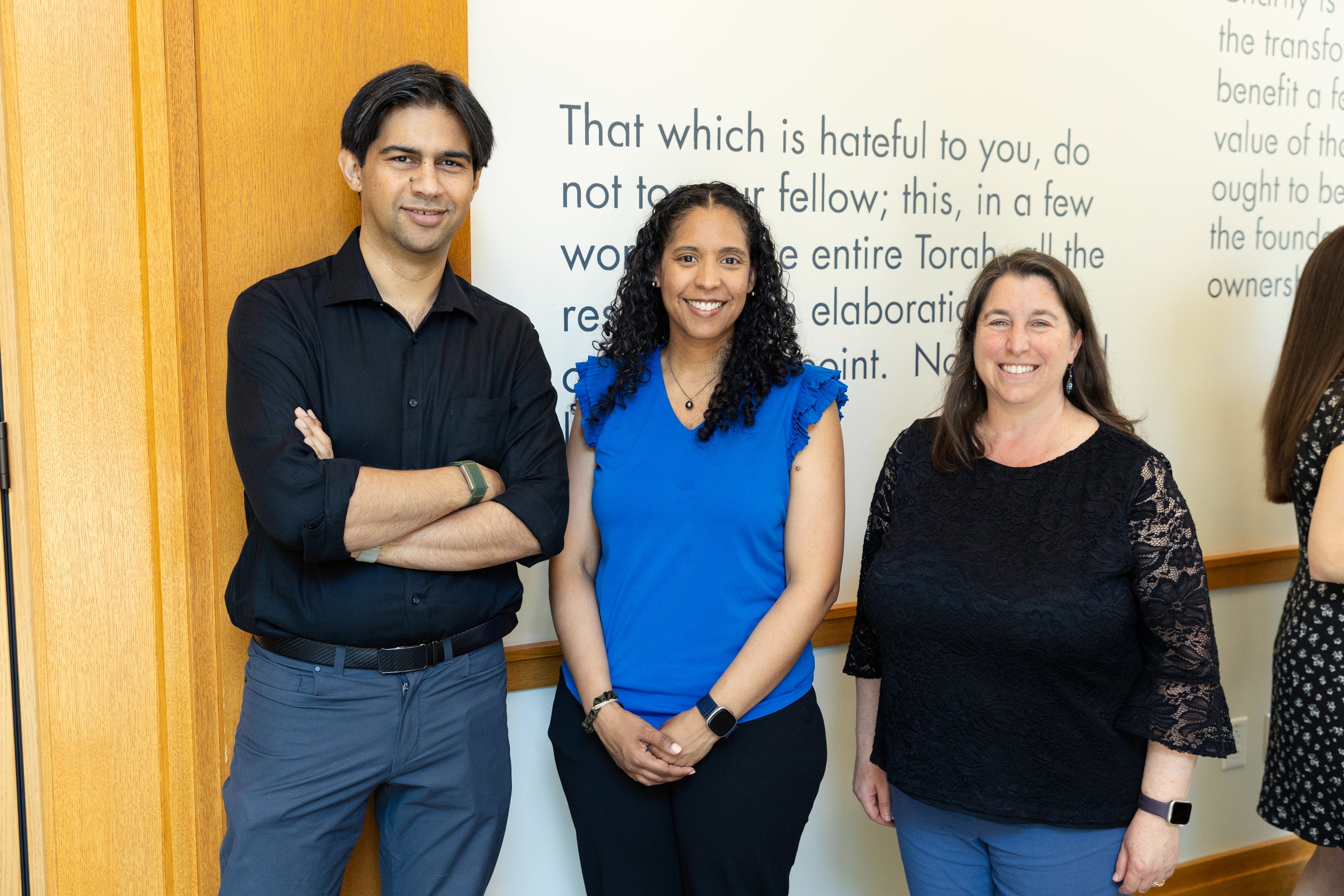 Three individuals stand together in front of a wall with text, smiling for the camera.
