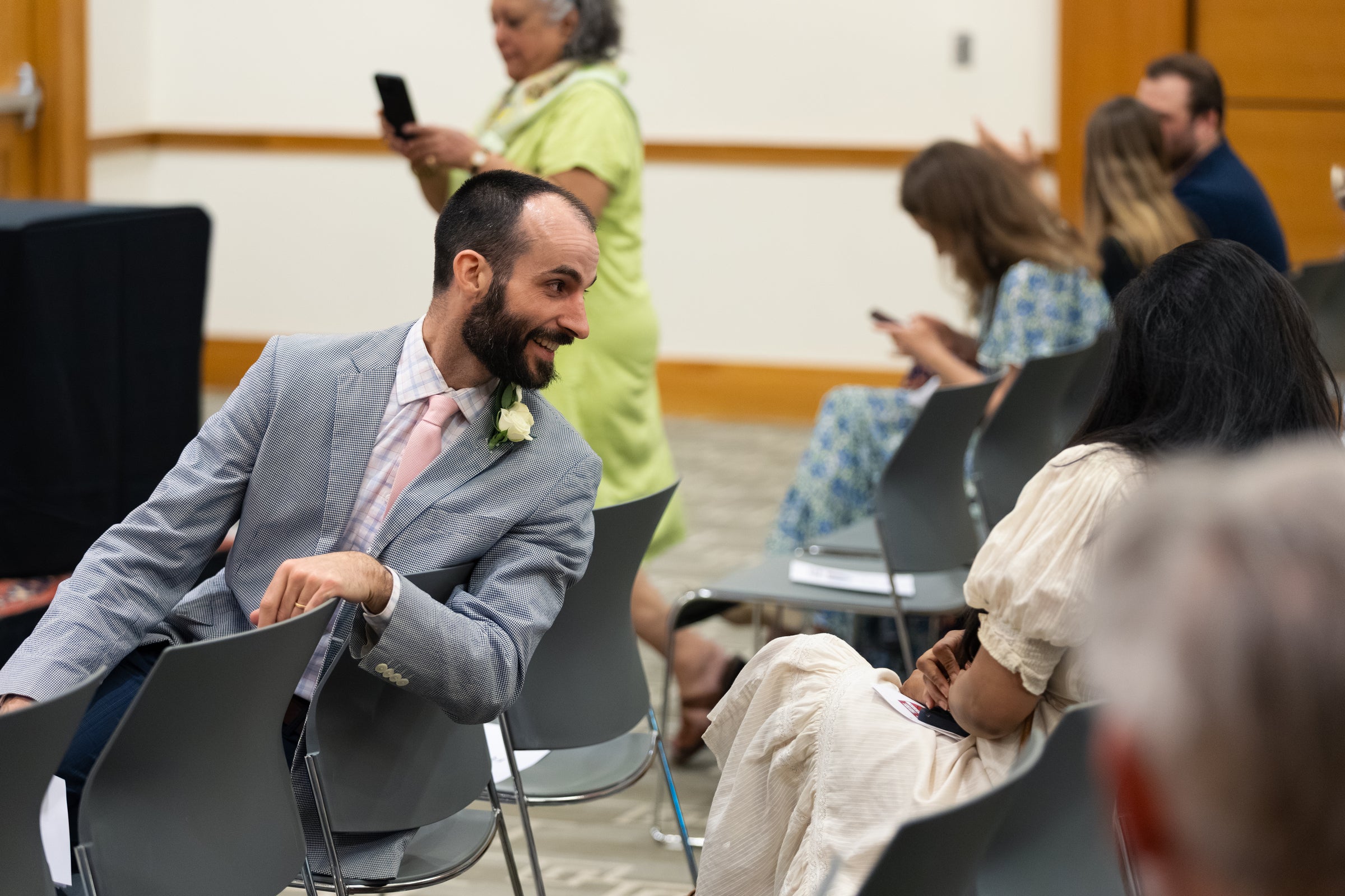A person in a light gray suit with a pink tie and a white flower boutonniere sits in an audience and turns around to talk to some seated in the row behind him.