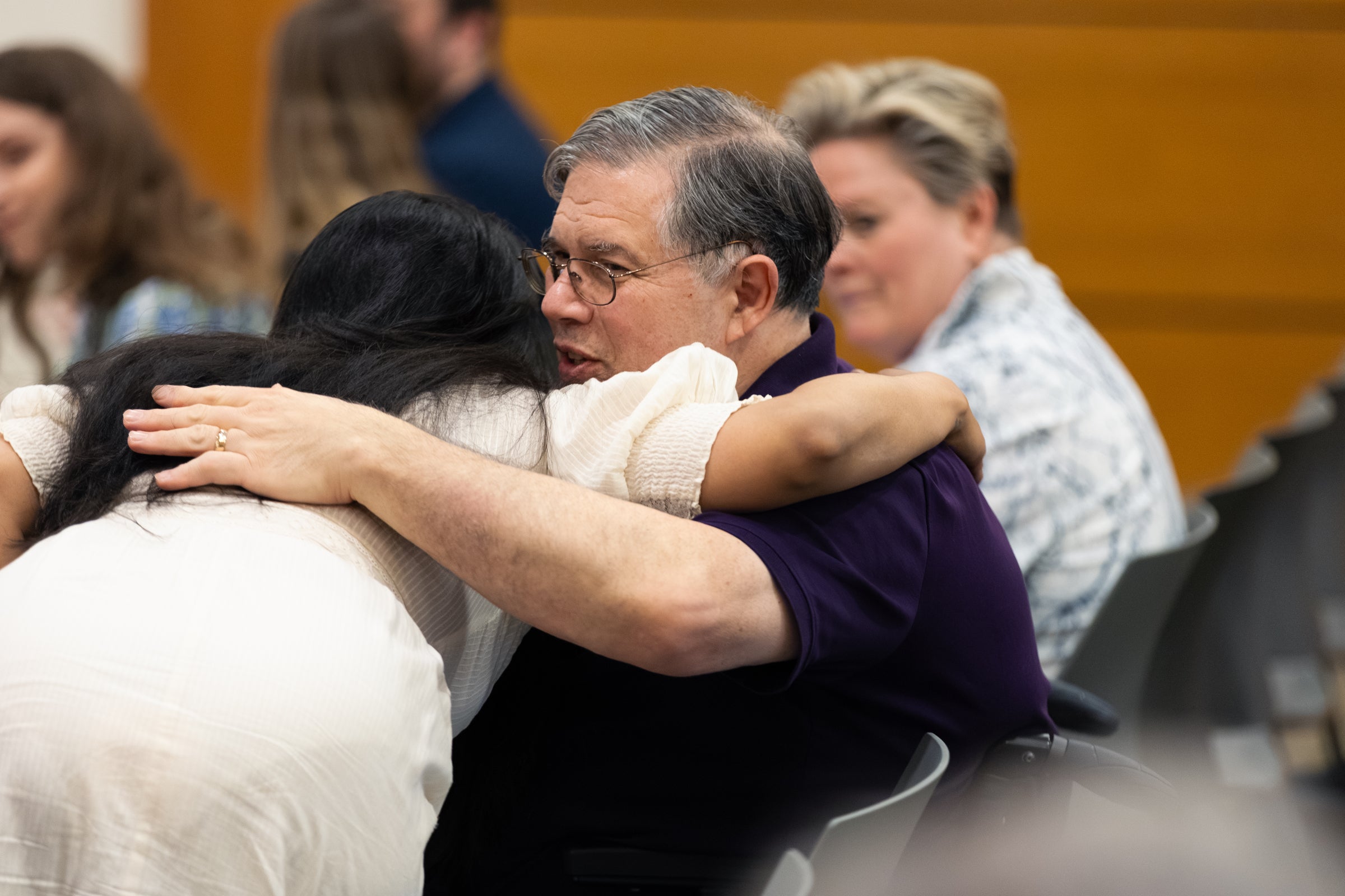 A person in a white dress bends down to hug a person in a black shirt that is seated.