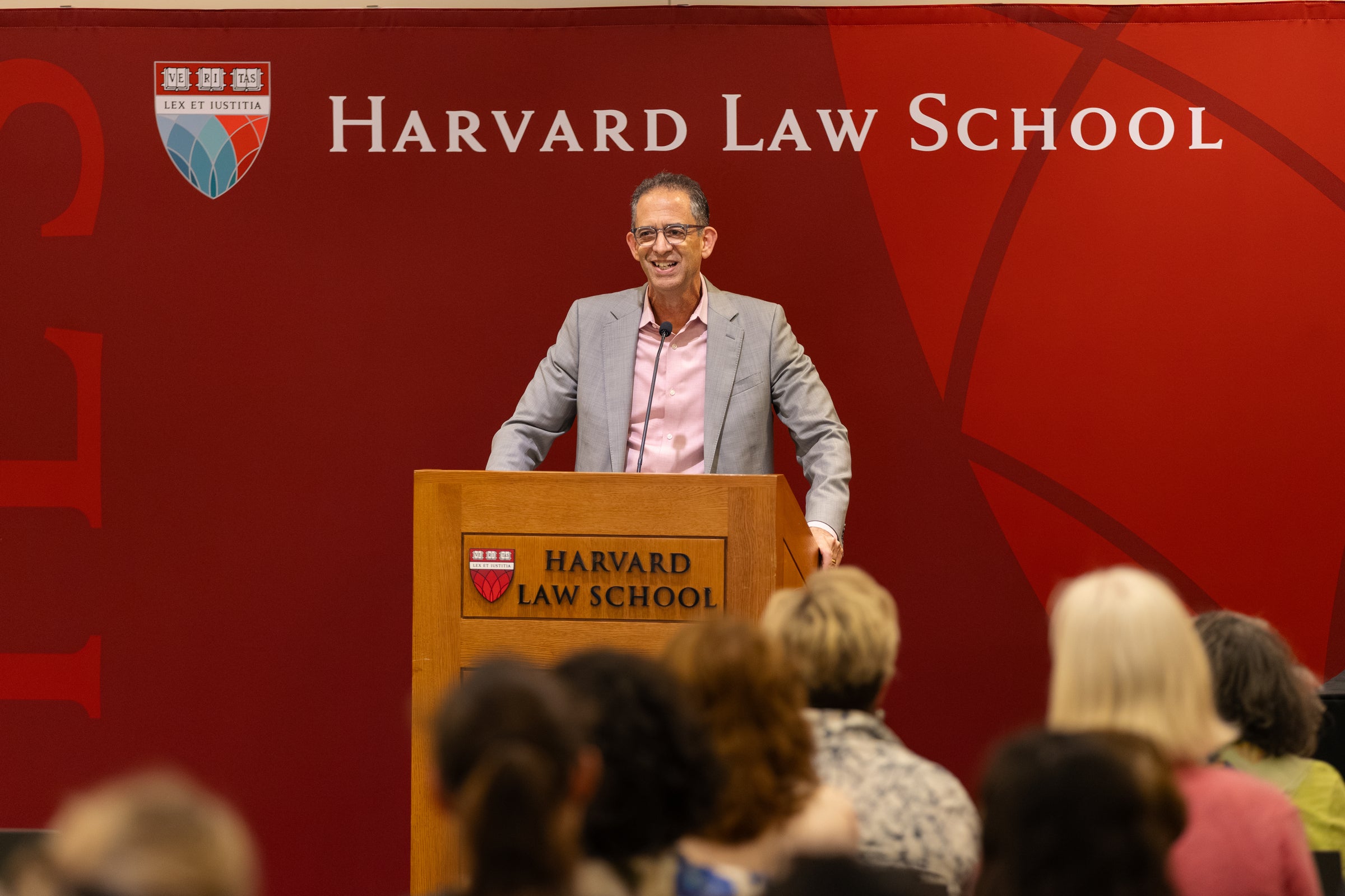 Dean Goldberg speaks at a podium to an audience, in front of a red backdrop displaying the shield and text 