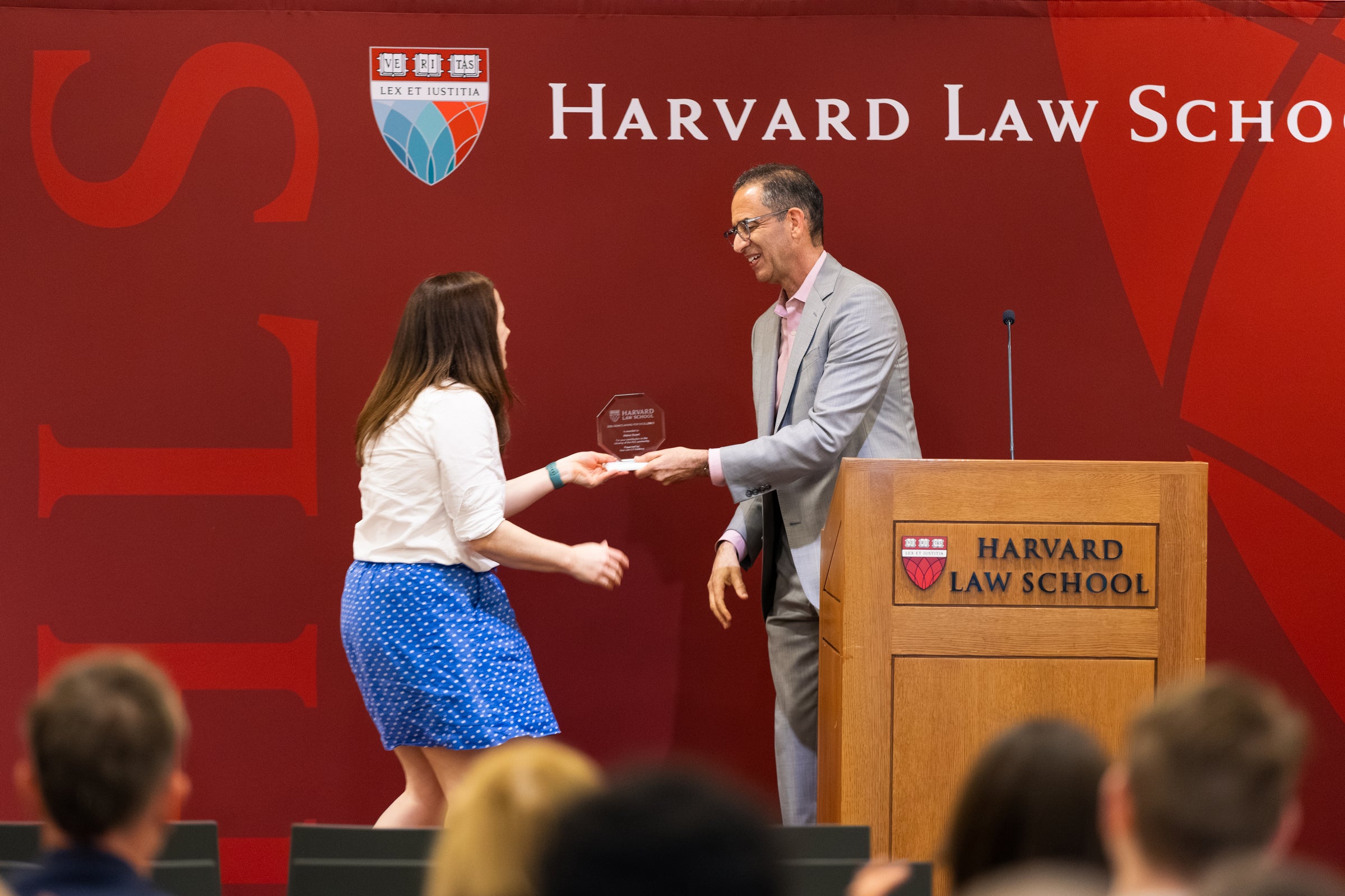 A person in a blue skirt and white shirt accepts an award from Dean Goldberg next to a podium.