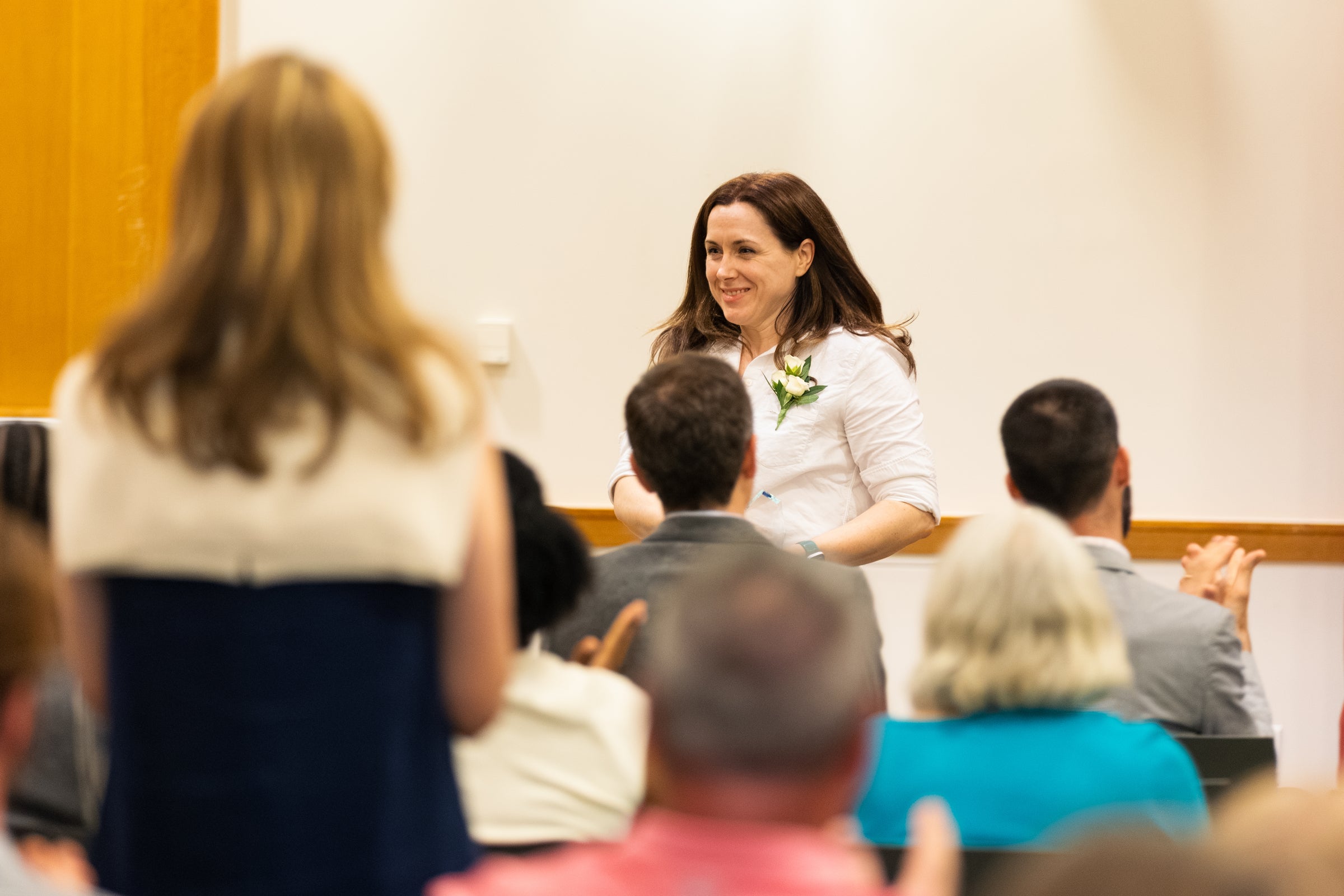 A person in a white shirt smiles to an applauding audience as she returns to her seat after accepting an award.