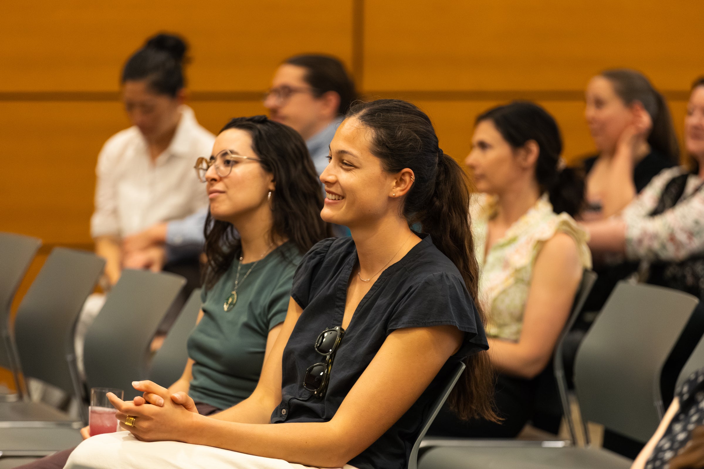 A group of people seated in an audience, with one individual smiling warmly while listening to a speaker.