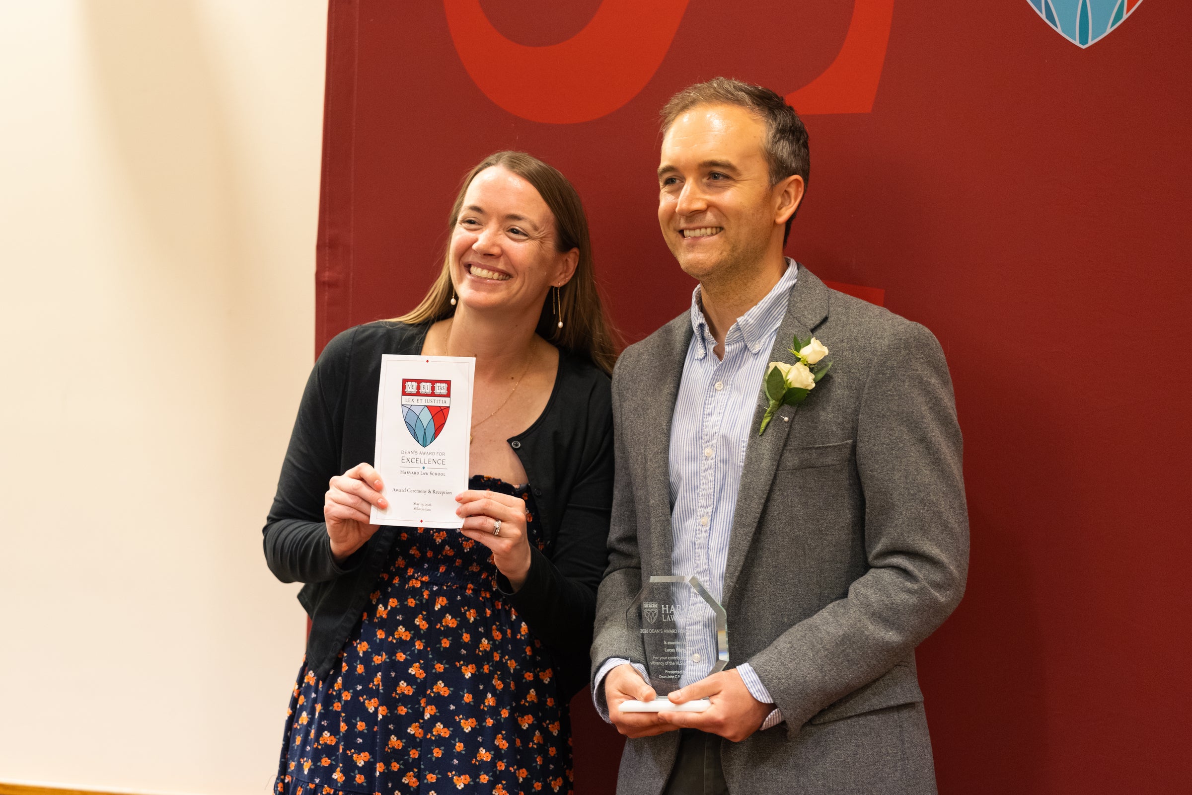 Two individuals stand together and smile for a photo, one holding an event program and the other holding an award, in front of a red backdrop.