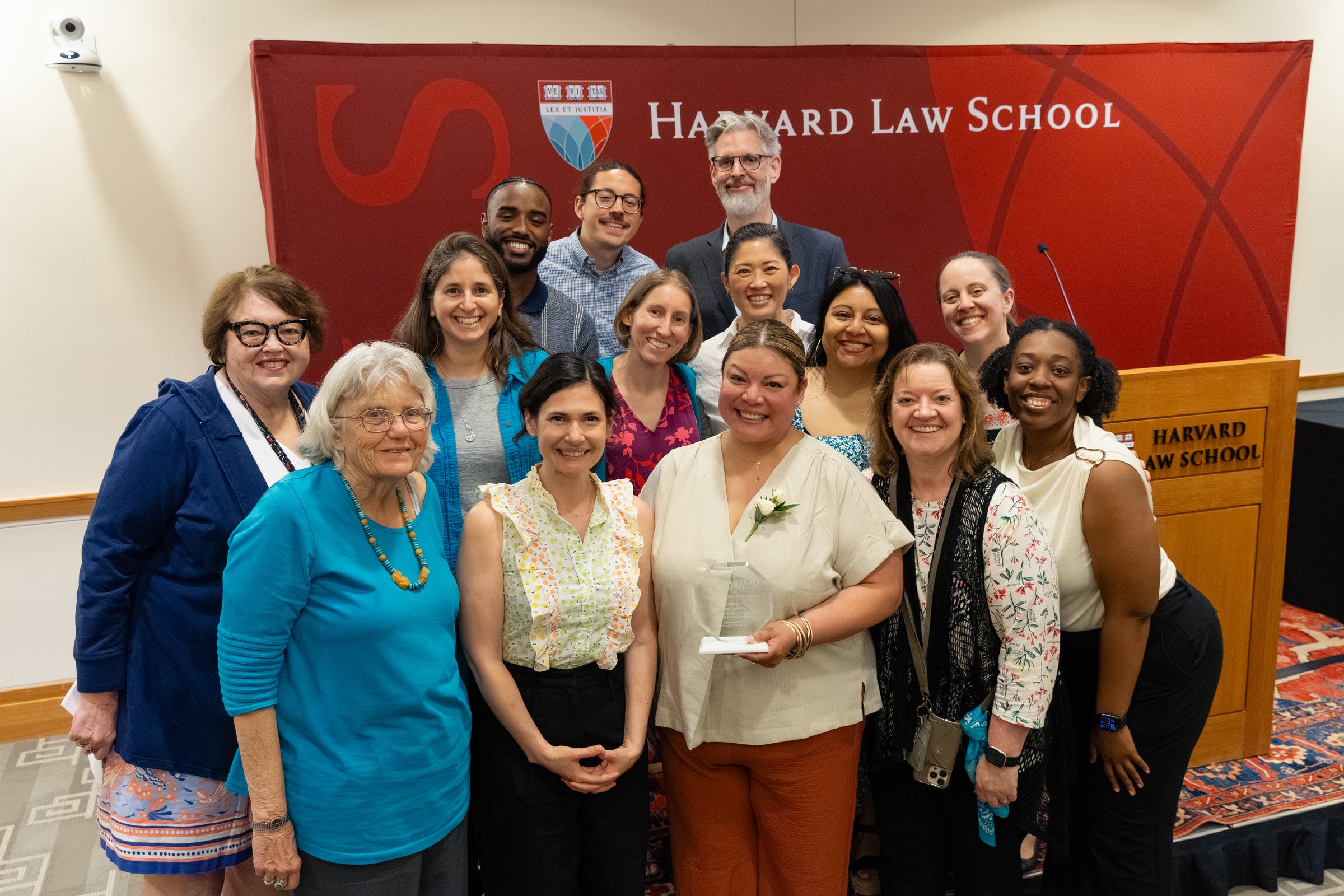 A group of people pose for a photo in front of a red Harvard Law School backdrop, with one individual in the front holding an award.