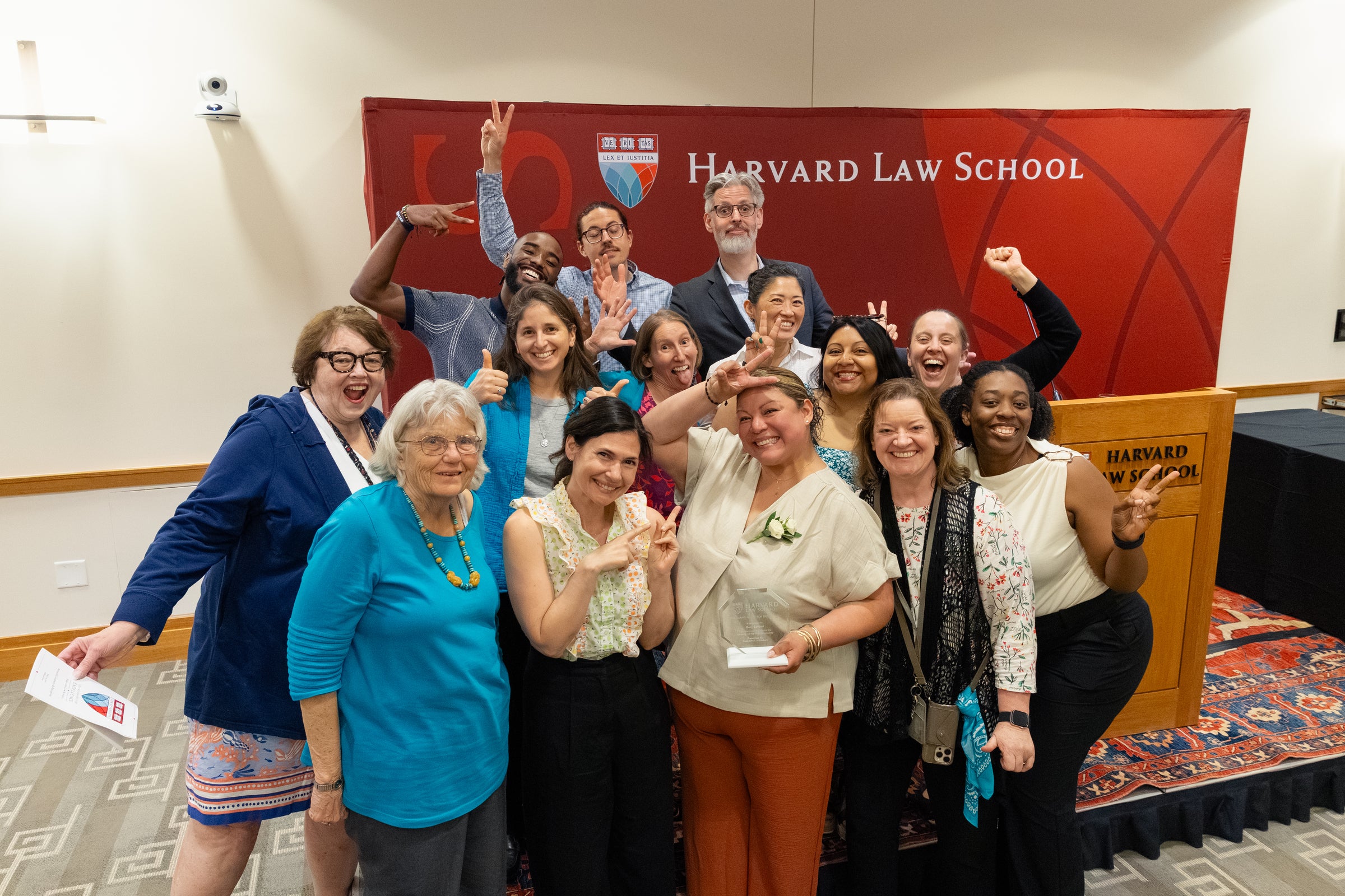 A group of people pose cheerfully in front of a Harvard Law School backdrop, with one person in front holding an award and everyone making celebratory gestures.