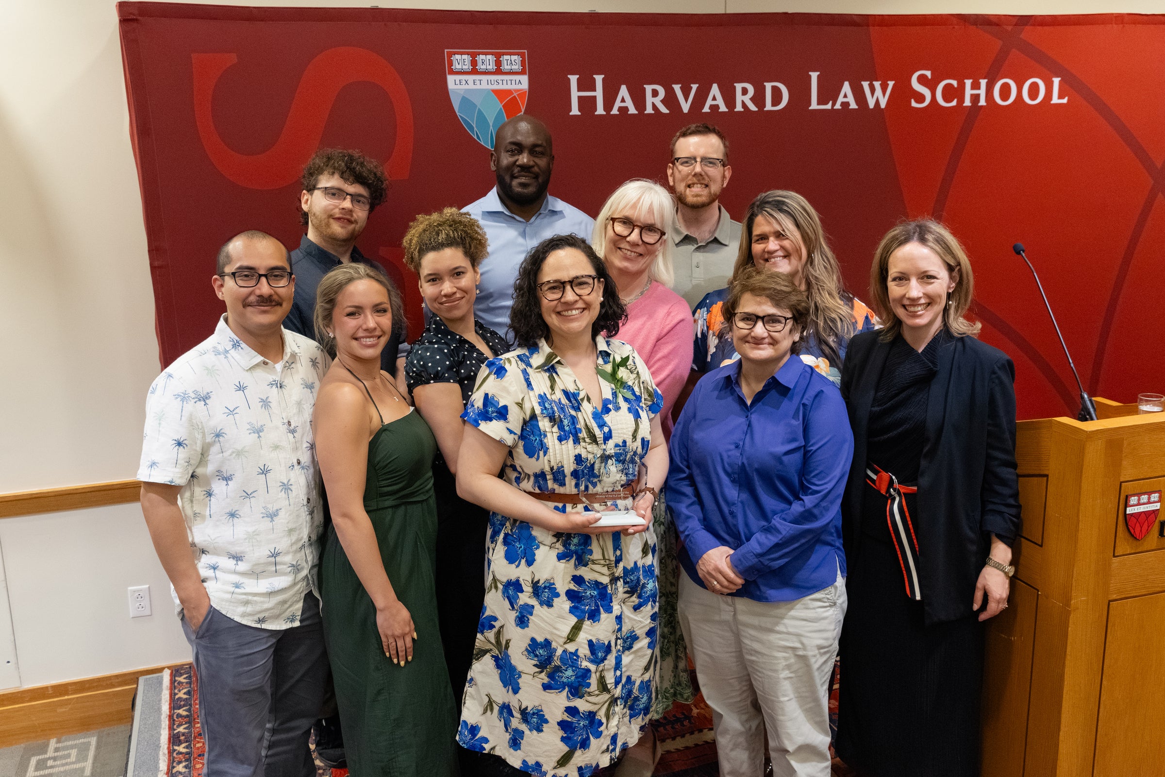 A group of eleven individuals pose for a photo in front of a Harvard Law School backdrop, with one person in the front holding an award.