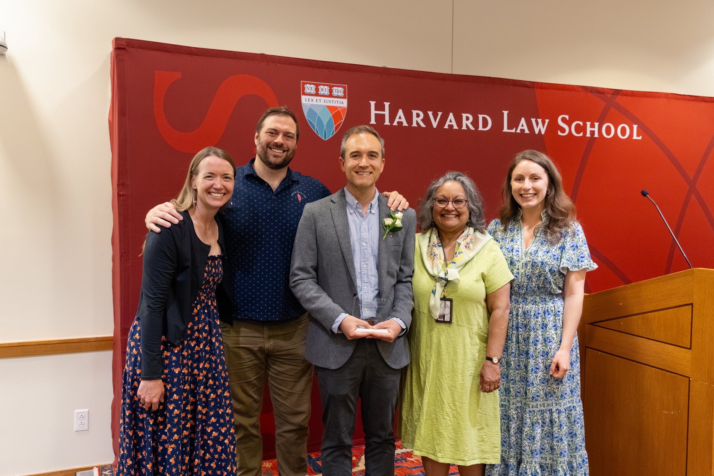 Five individuals pose together in front of a Harvard Law School backdrop and smile for the camera.
