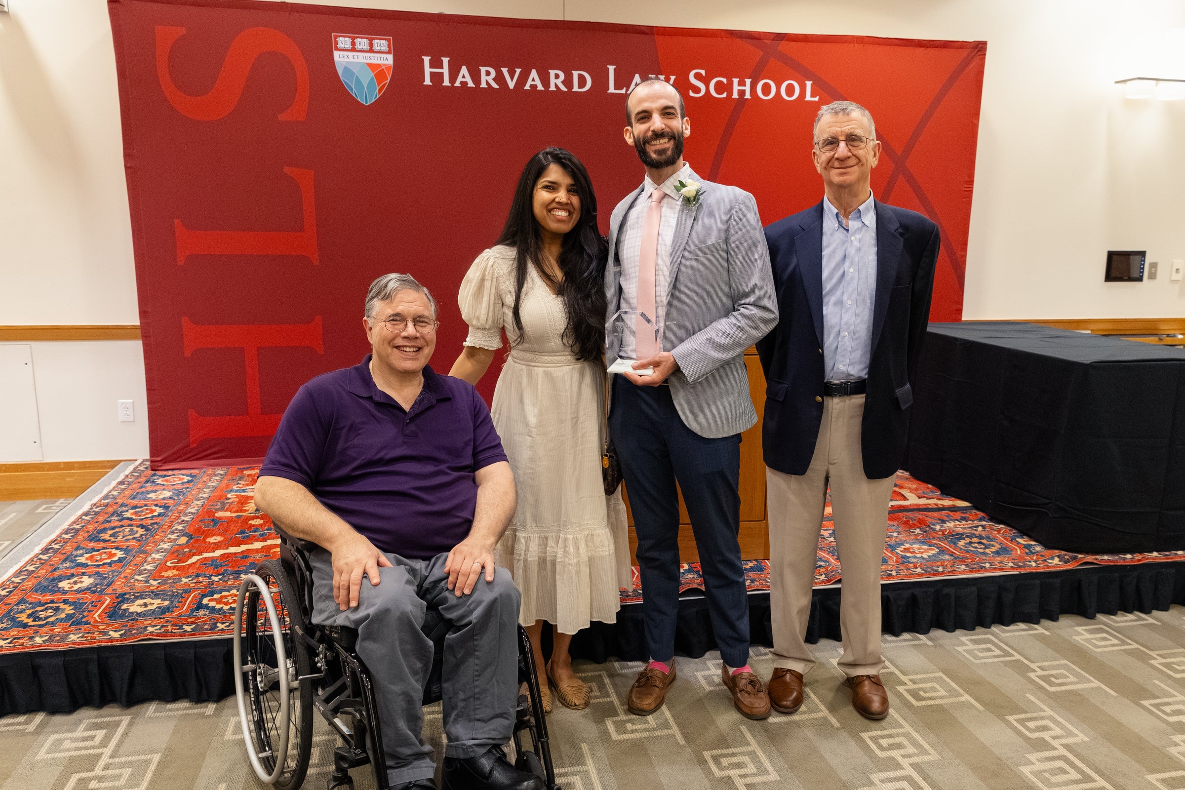 Four individuals pose for a photo in front of a stage and a Harvard Law School backdrop, with one person in the center holding an award and another seated in a wheelchair.
