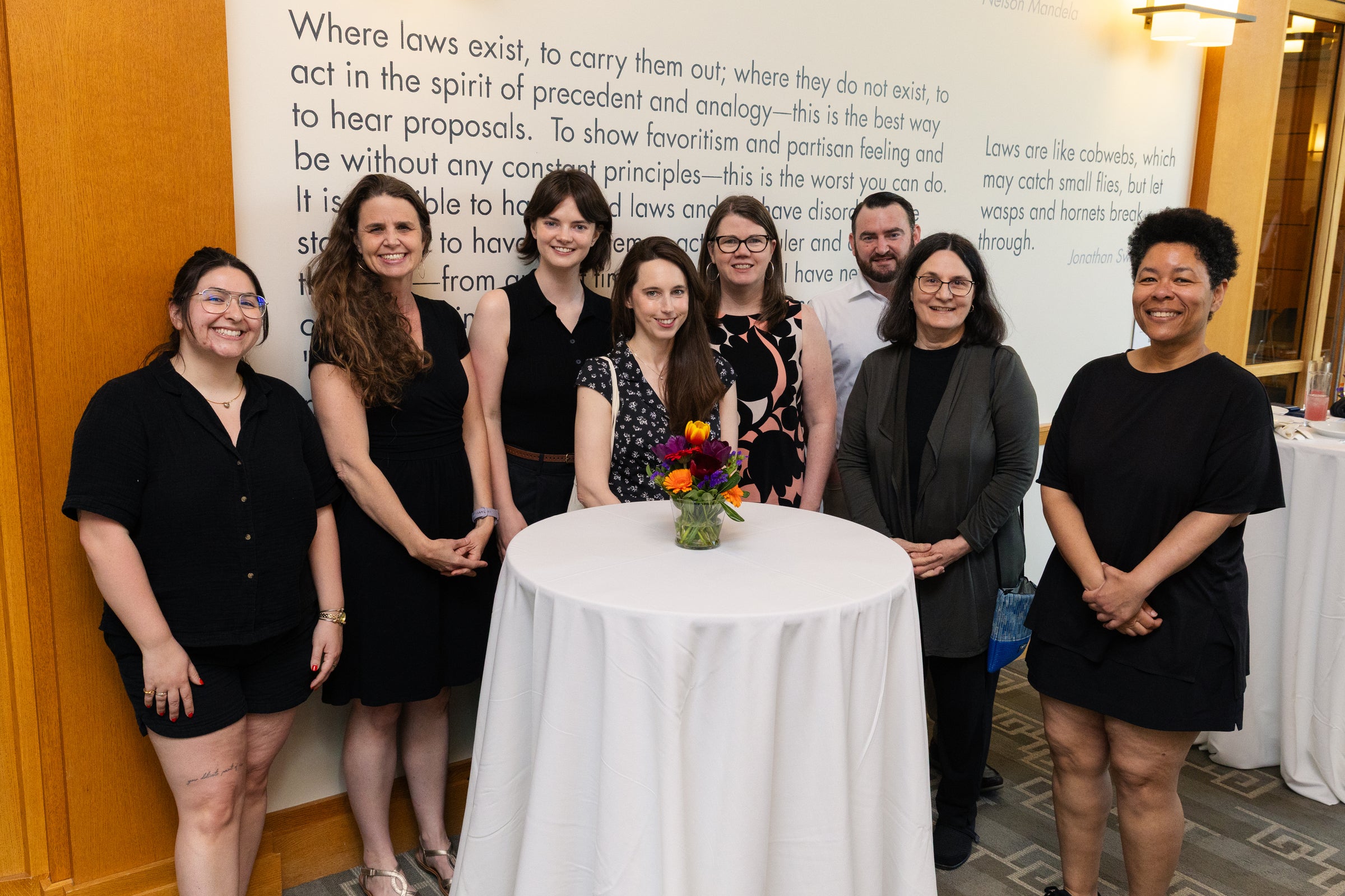 A group of eight individuals poses together in front of a wall with text, standing around a small round table with a floral centerpiece.