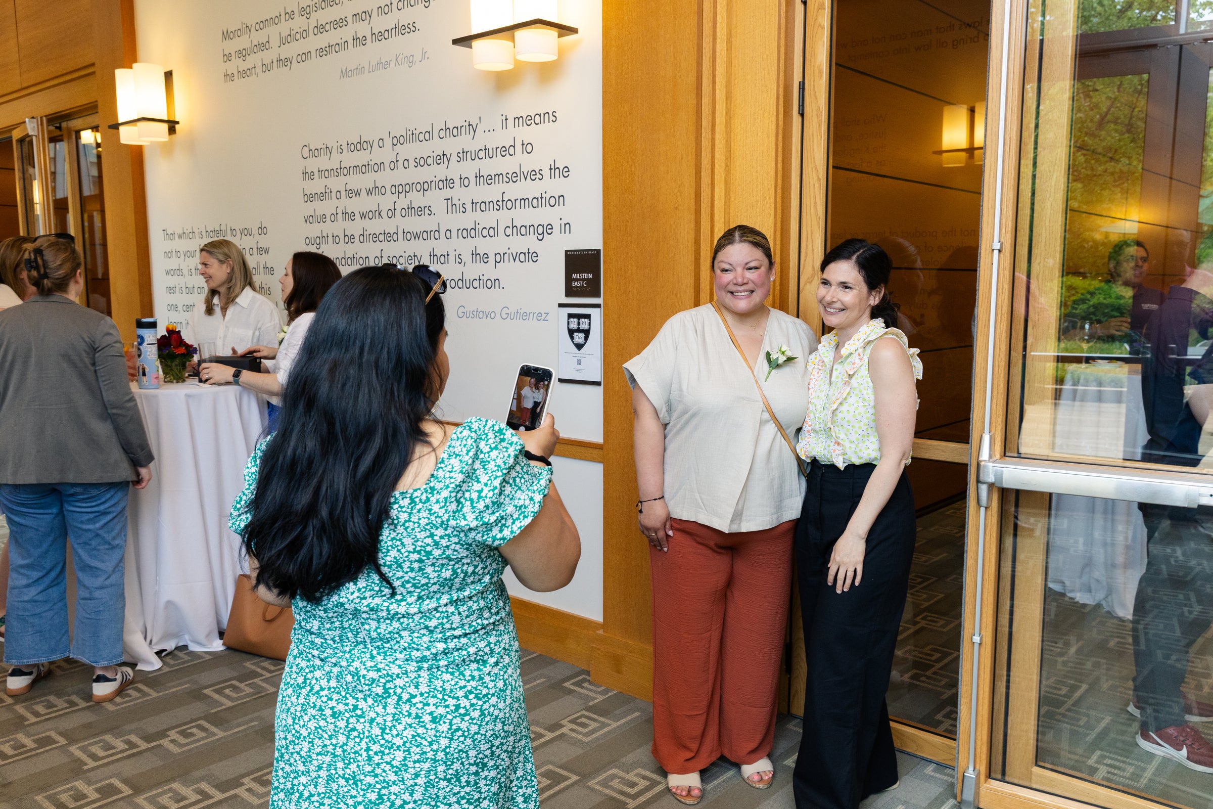A person takes a photo of two individuals smiling together near a doorway, with others socializing in the background.