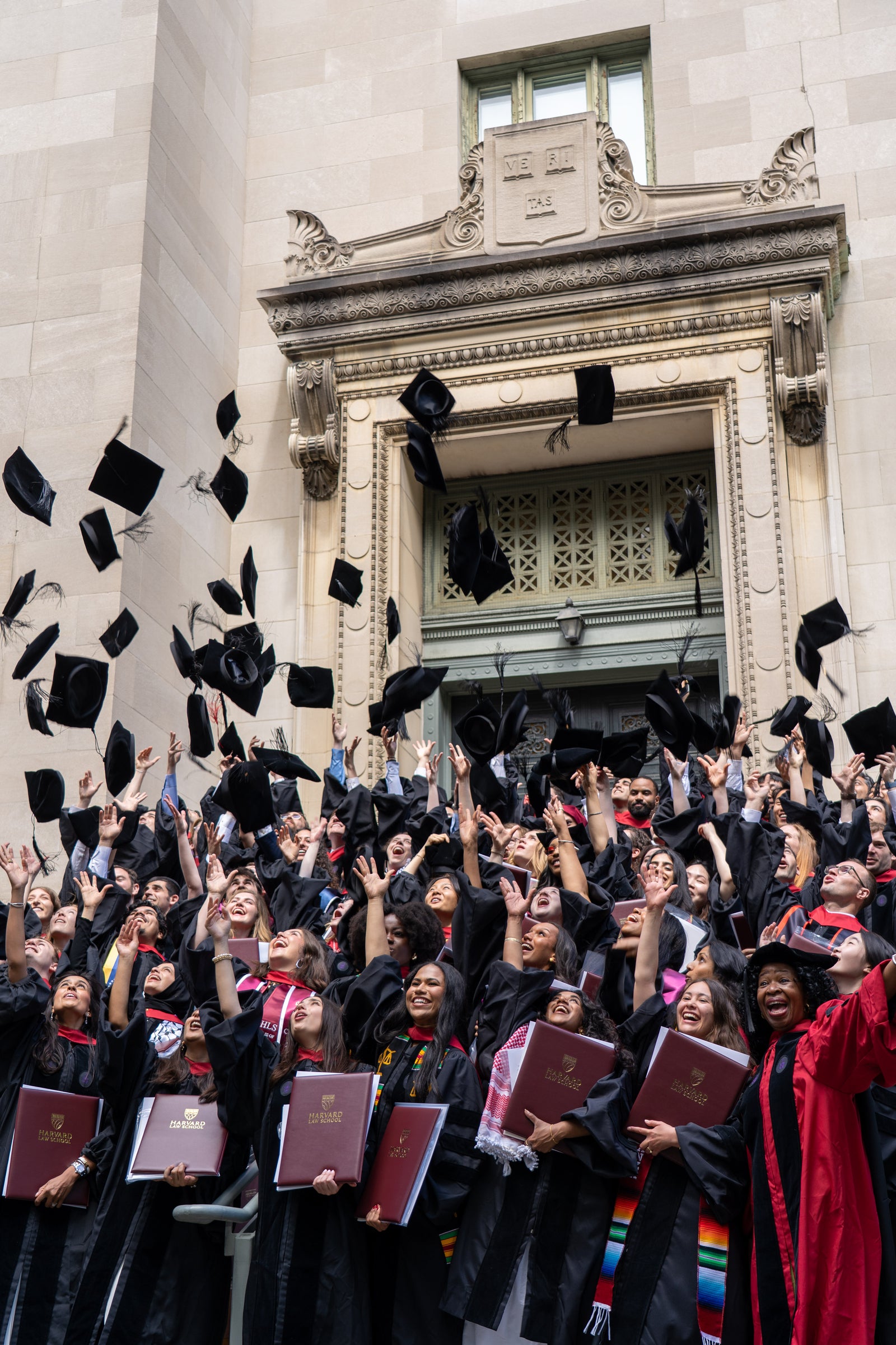 Graduates in academic regalia throw their caps into the air while posing on stairs for a group photo.