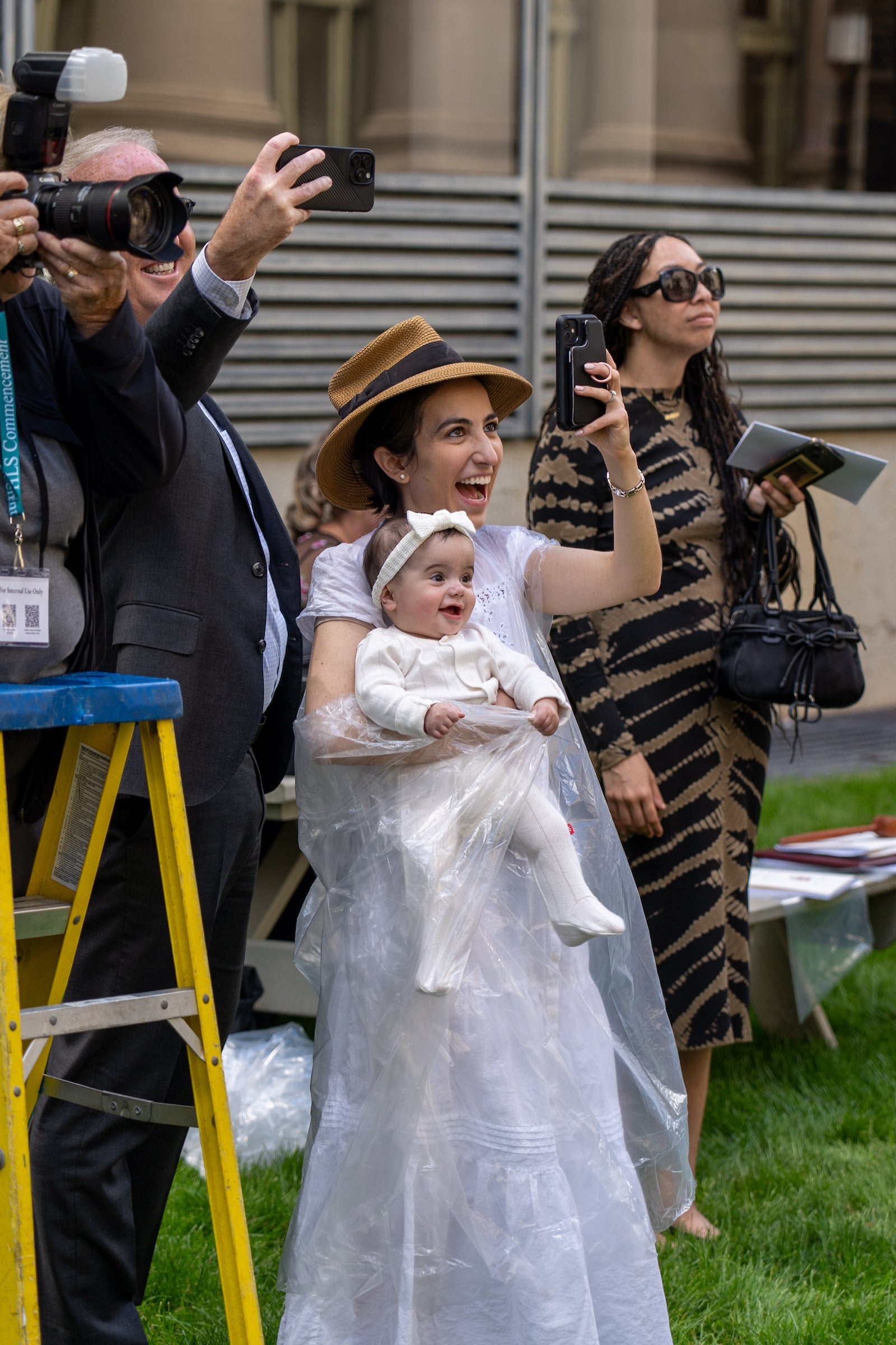 An attendee at commencement wearing a rain poncho holding her baby while taking a photo.