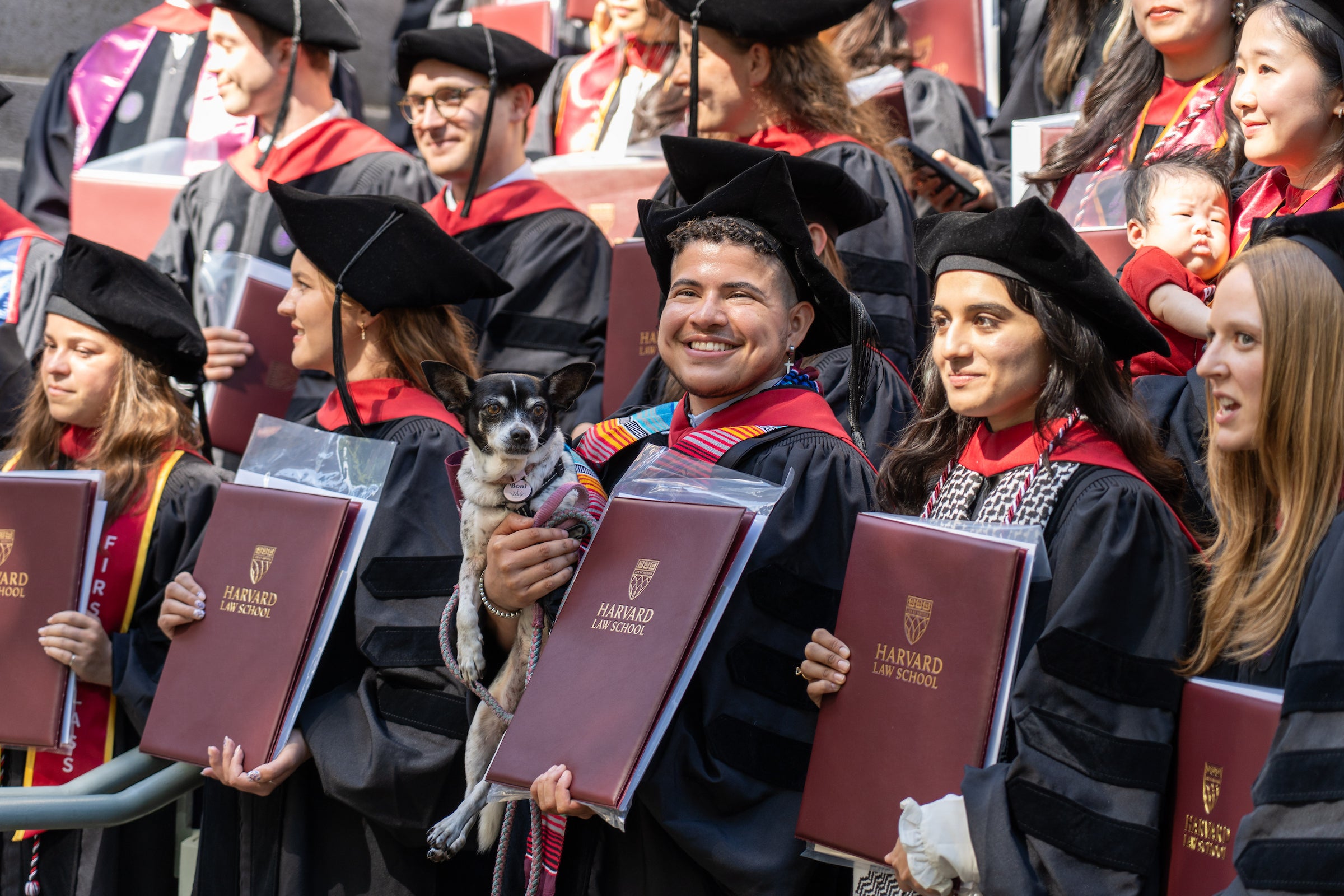 A group shot of students posing for a group shot holding diplomas with a student in the front row smiling with his dog.