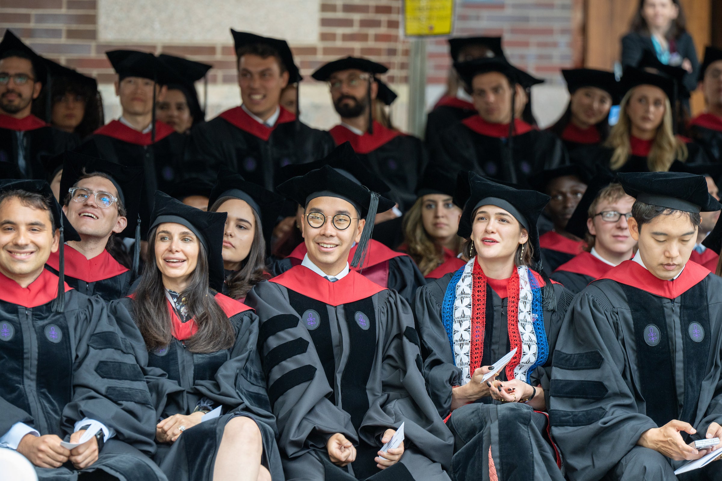 Rows of graduate students in commencement regalia sitting under a tent.