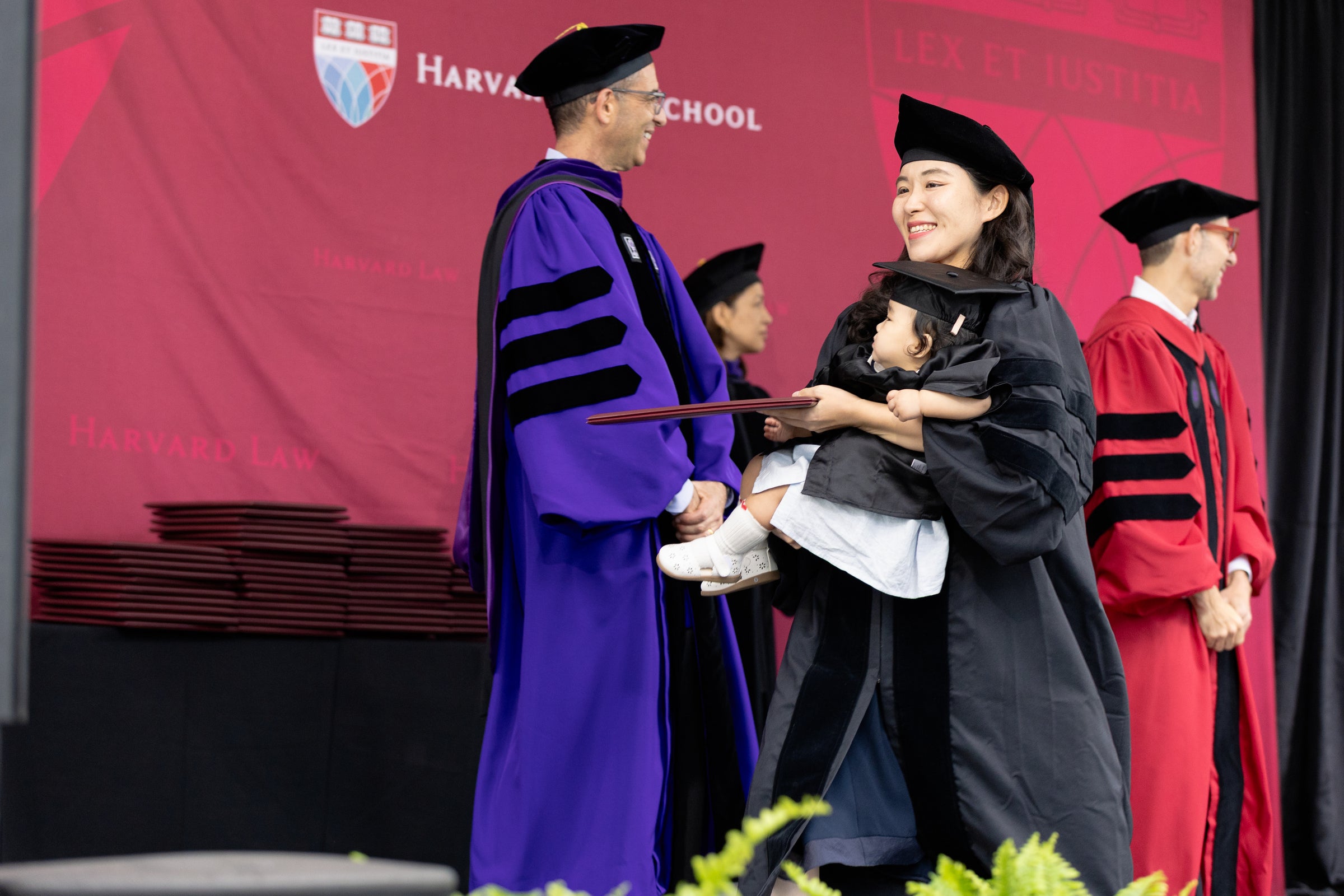 Woman in Commencement regalia holds a young child on the stage as she gets her diploma from the dean.