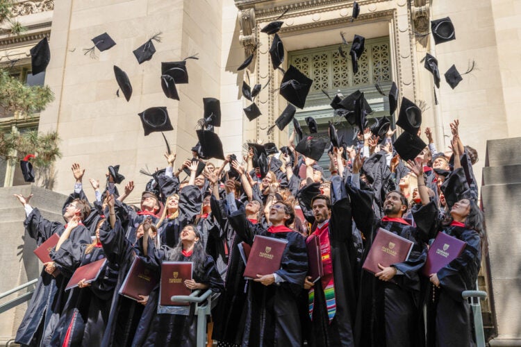 Students in graduation attire throw their caps into their air in front of an ornate stone building.