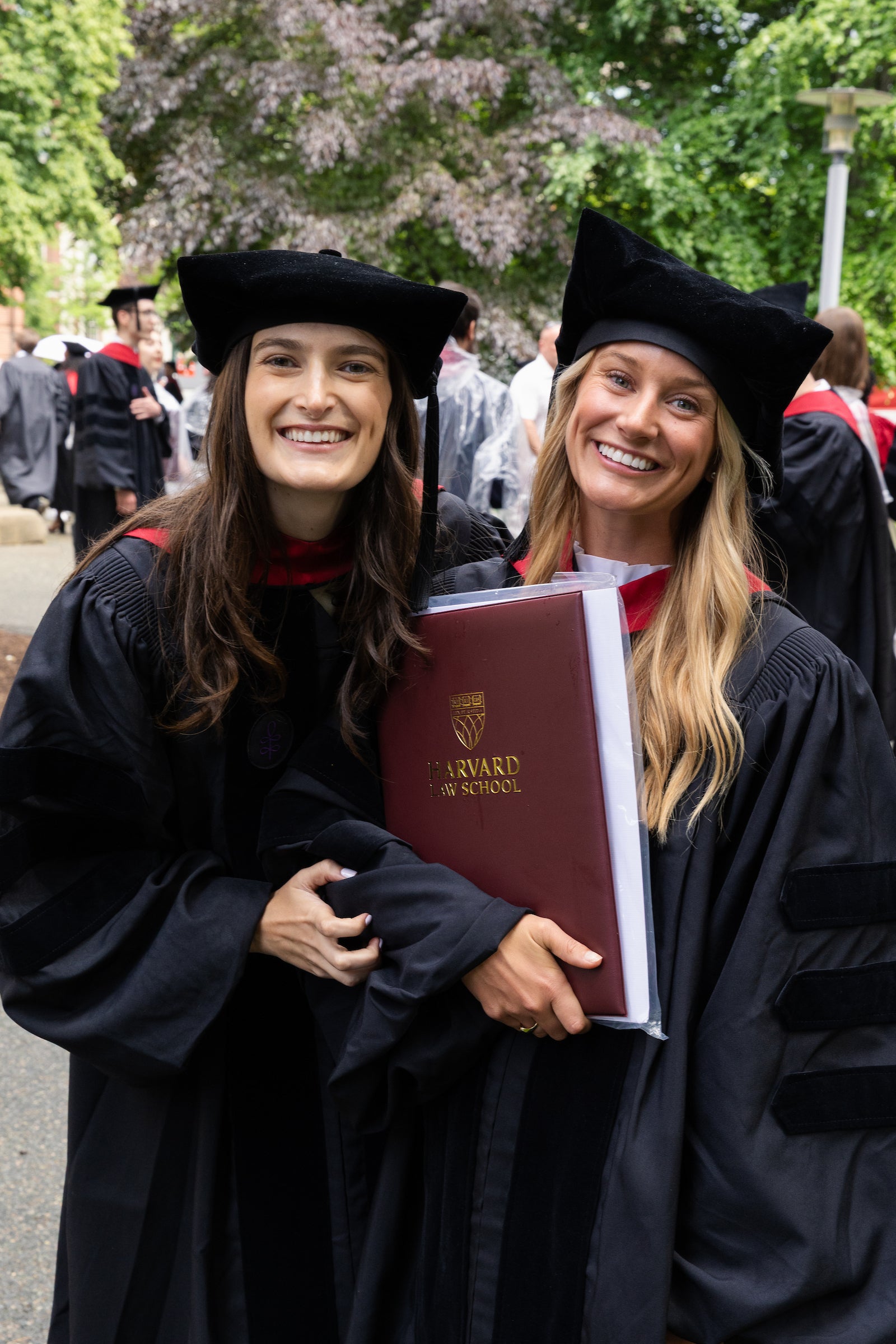 Two graduates in black caps and gowns smile while holding a Harvard Law School diploma folder outdoors.