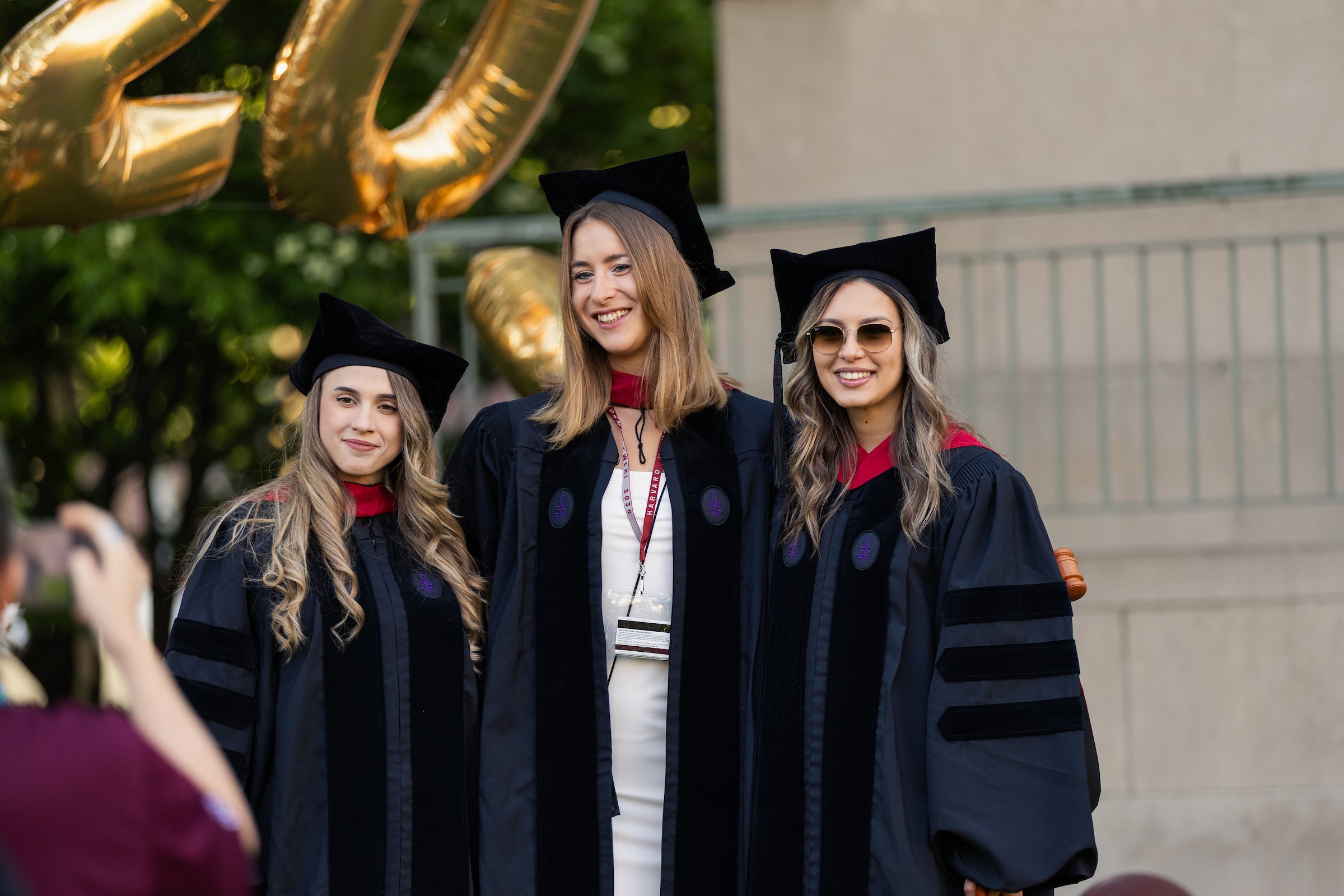 Three graduates in regalia smile for the camera outdoors, with large gold balloons visible in the background.
