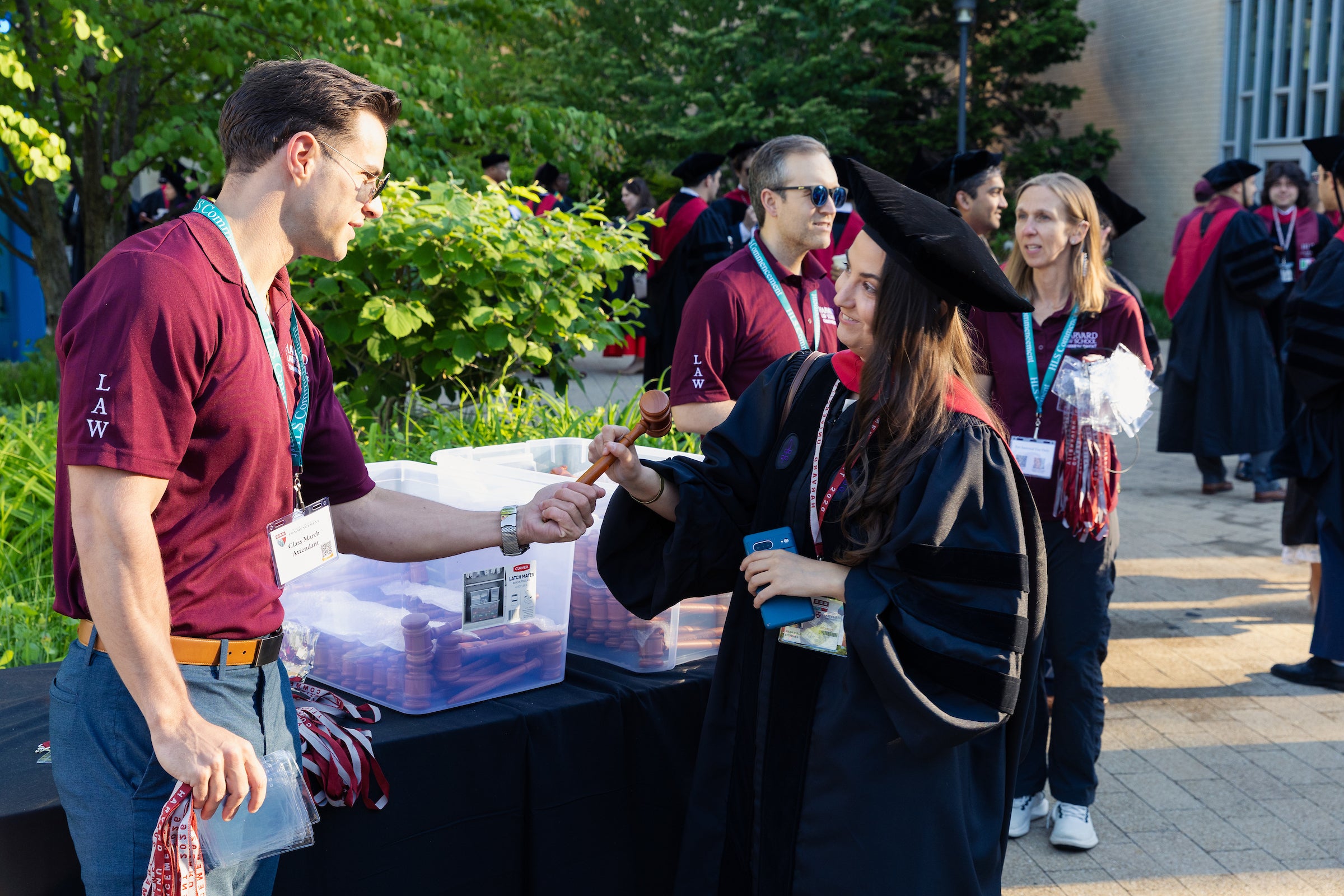 A graduate in regalia receives a wooden gavel from a volunteer at an outdoor event.