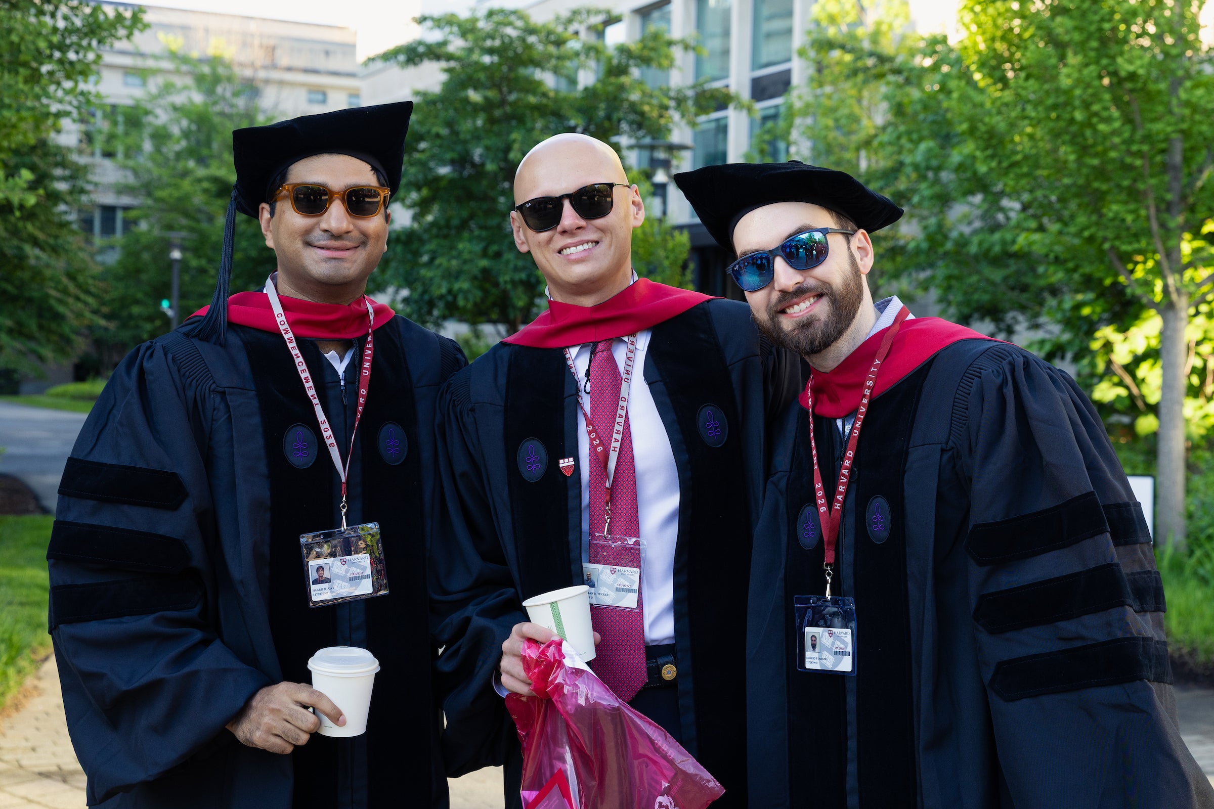 Three individuals in academic regalia pose outdoors, smiling while holding drinks and a plastic bag.