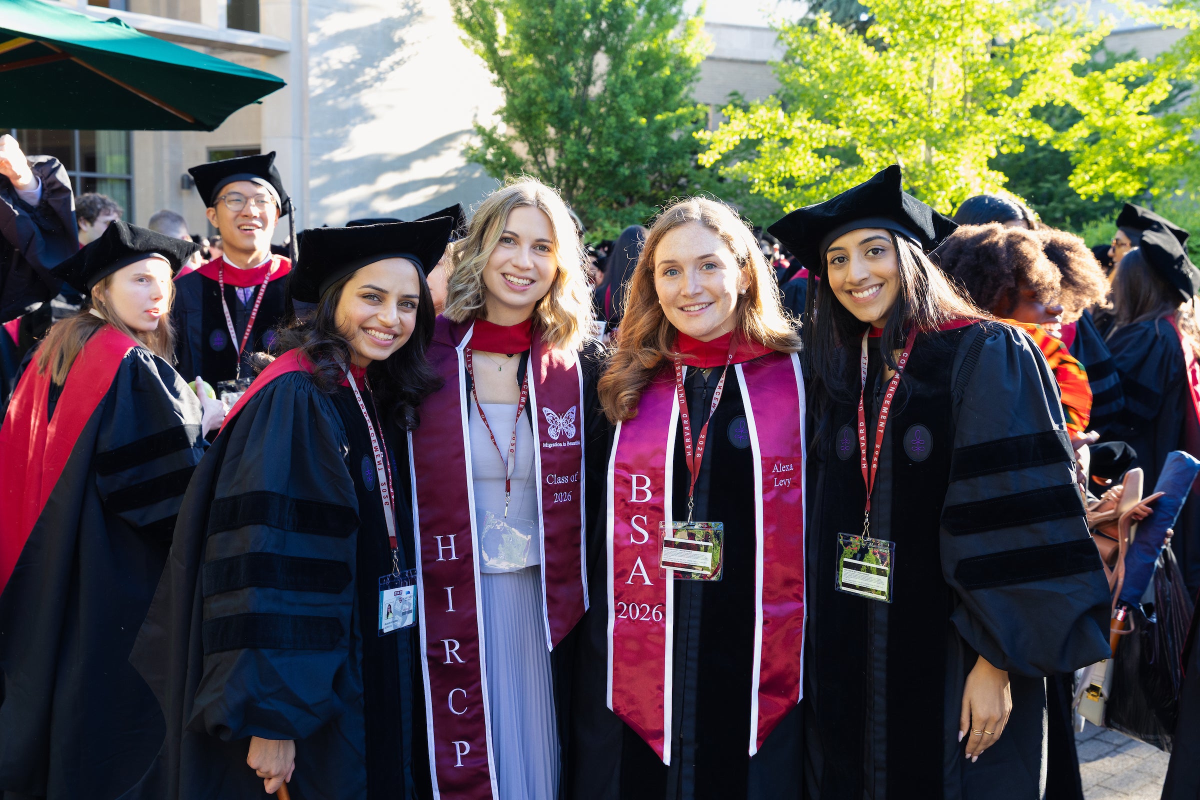 A group of graduates in black caps and gowns with maroon sashes stand together and smile for the camera in an outdoor courtyard.