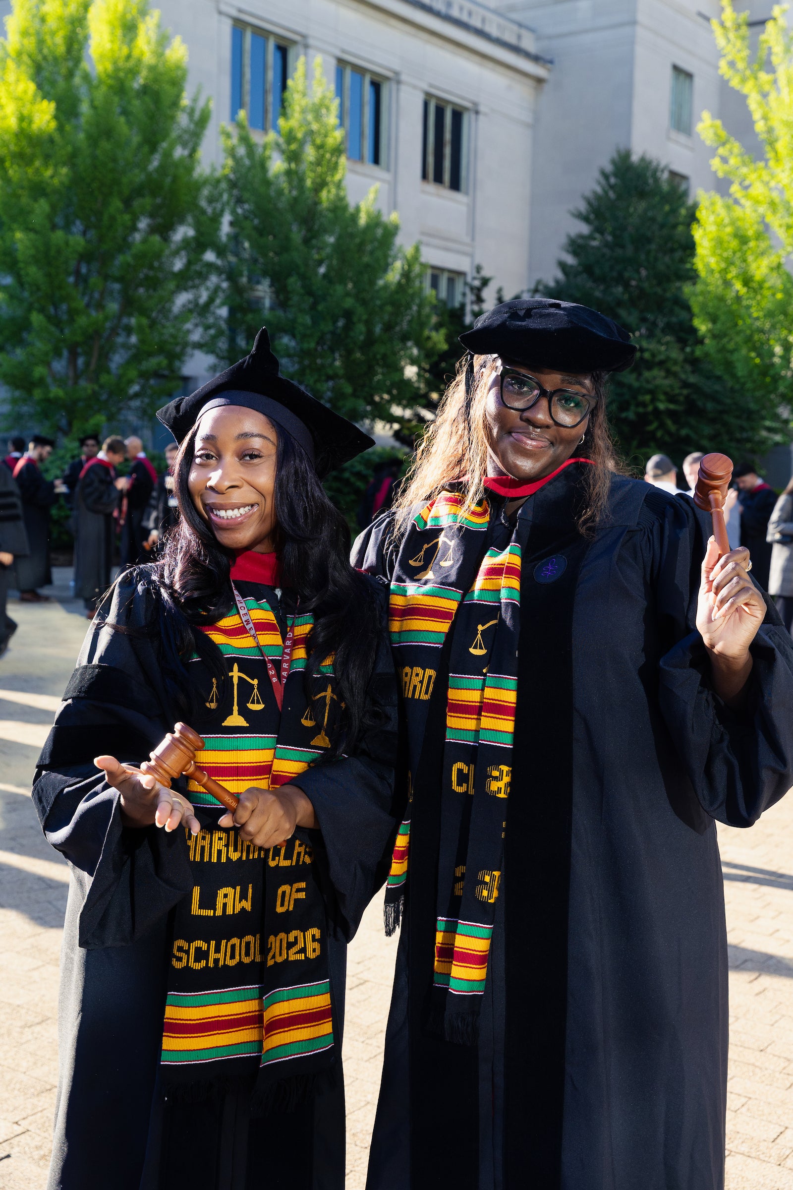 Two graduates in regalia pose for a photo outdoors while holding up gavels.