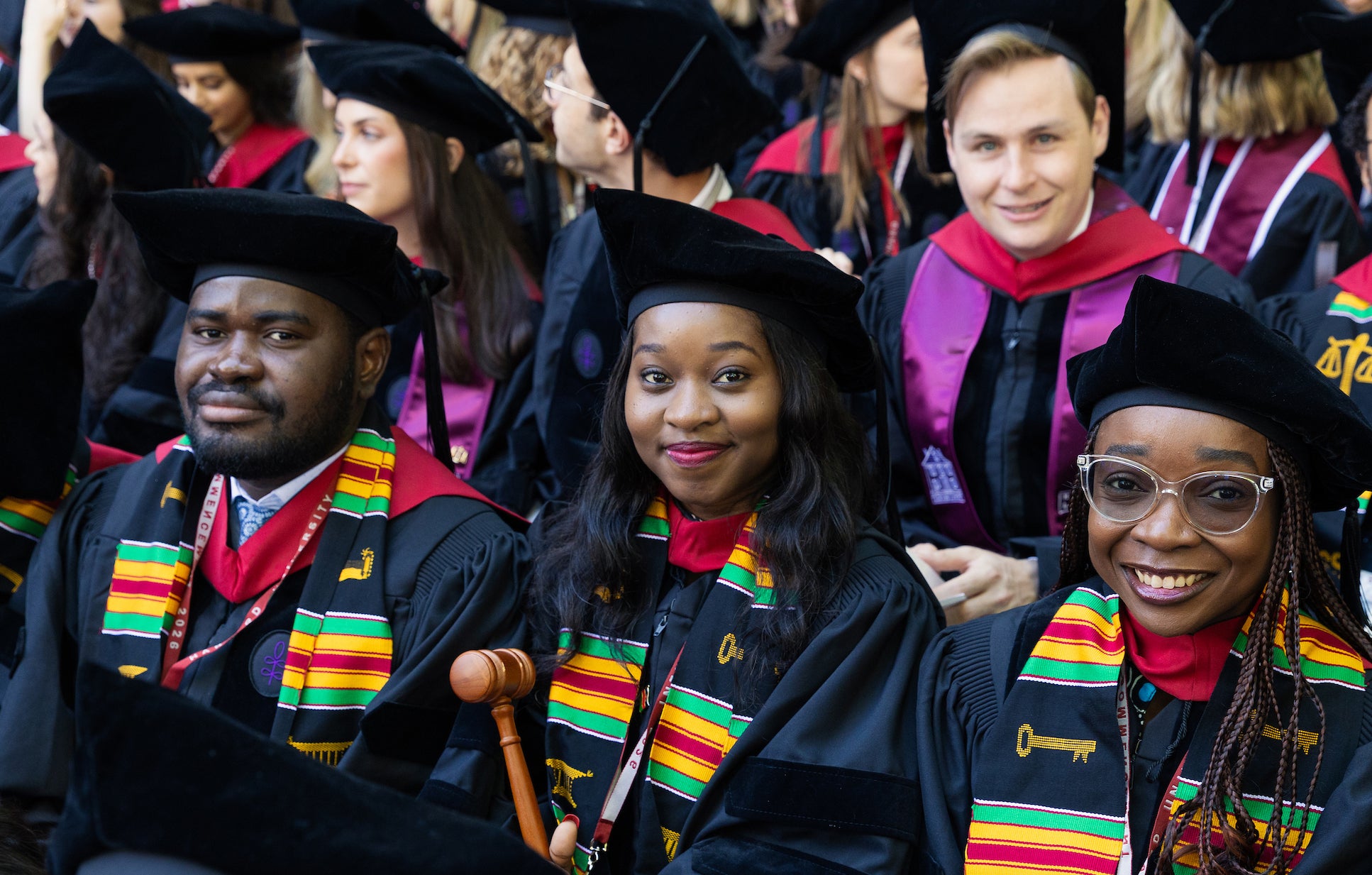 A group of graduates in regalia smile for the camera while seated for the ceremony.