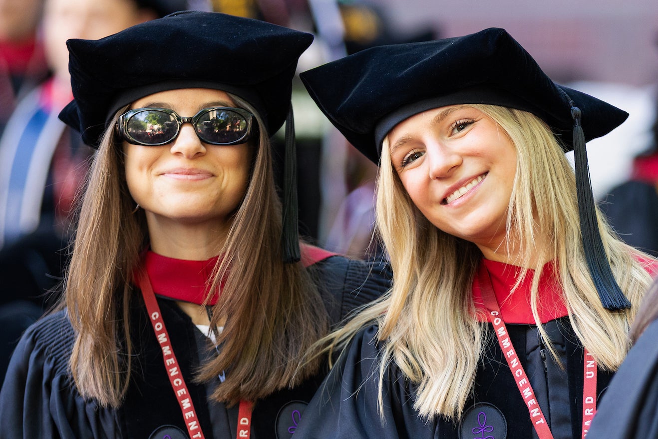 Two graduates in black caps and gowns with red lanyards smile at the camera during a Harvard commencement ceremony.