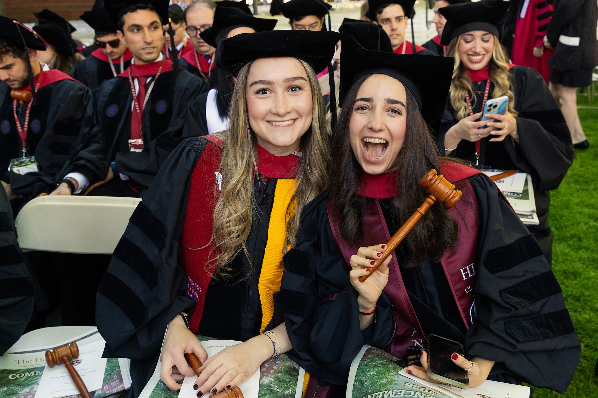 Two graduates in regalia smile for the camera while seated for commencement, one holding a wooden gavel.