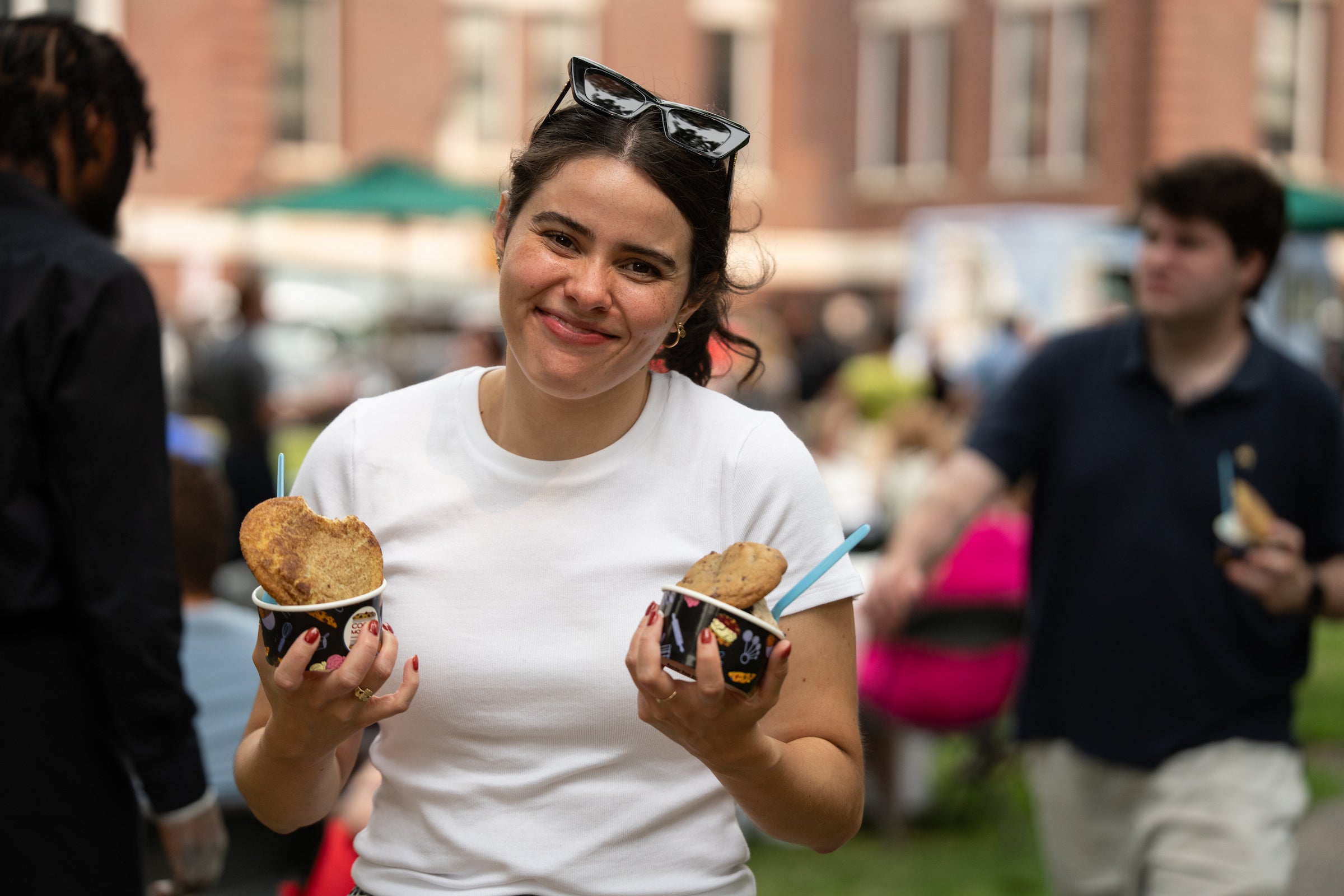 A person smiles while holding two cups of cookies and ice cream at an outdoor event.