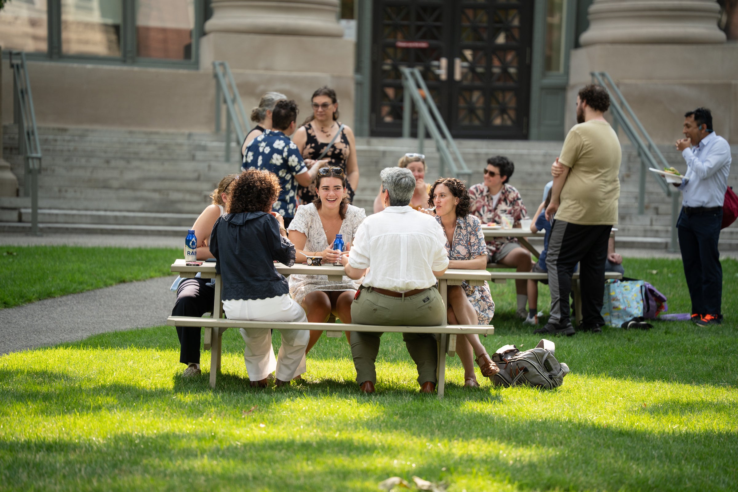 A group of people socialize and eat outdoors on a sunny day at a table on a grassy lawn in front of a building with columns.