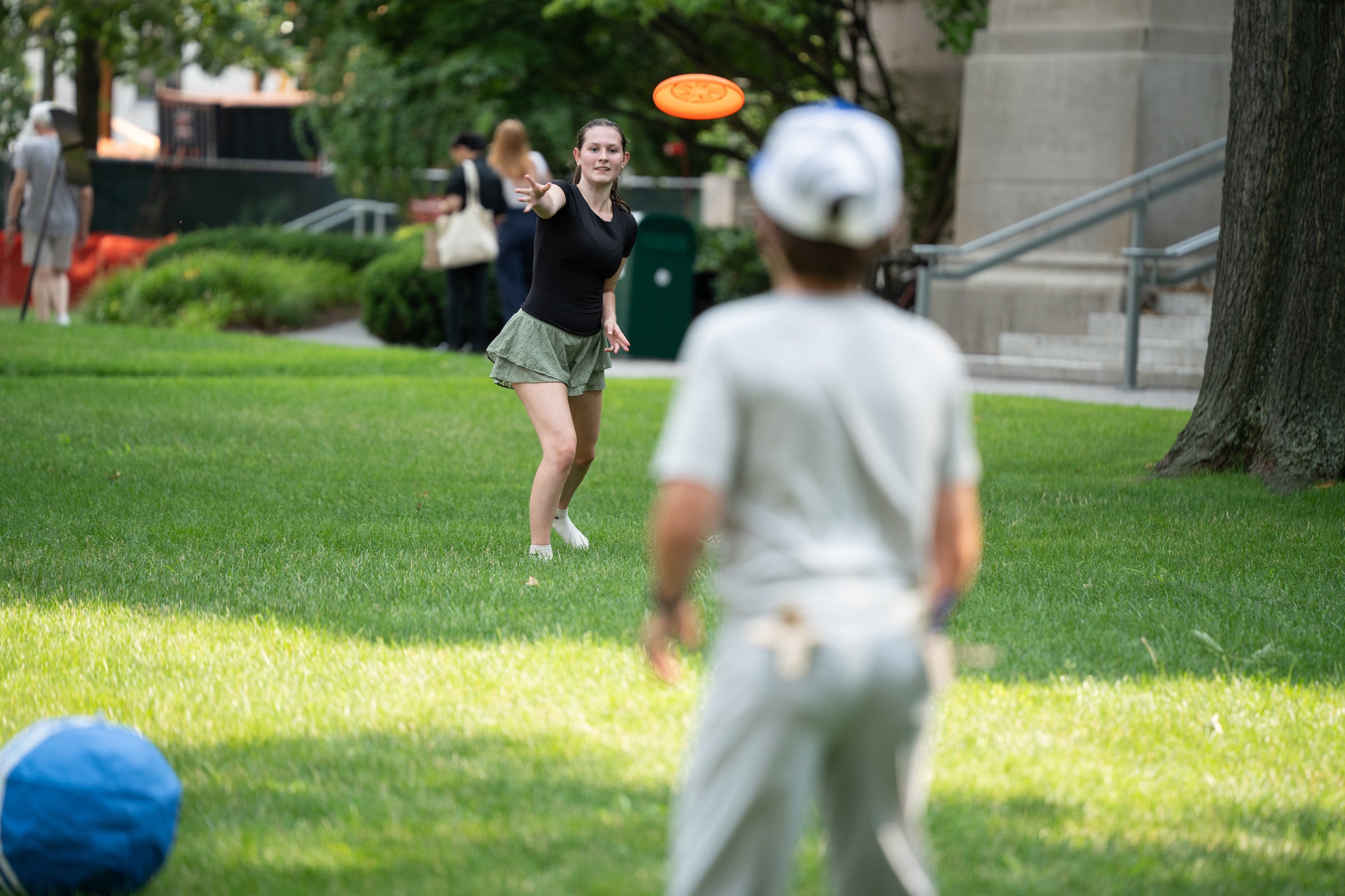 Two people play frisbee on a sunny grassy lawn, with one throwing the disc and another preparing to catch it.