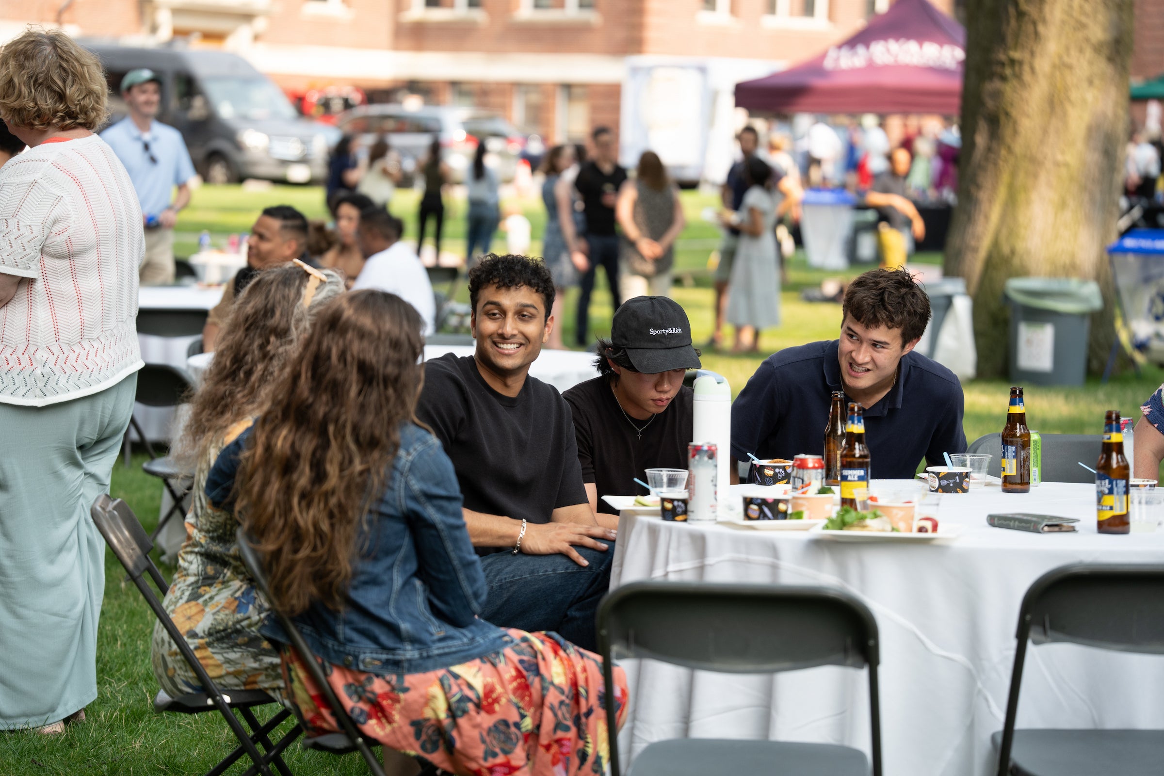 A group of young people socialize outdoors at a table with food and drinks, surrounded by other attendees and a grassy area.