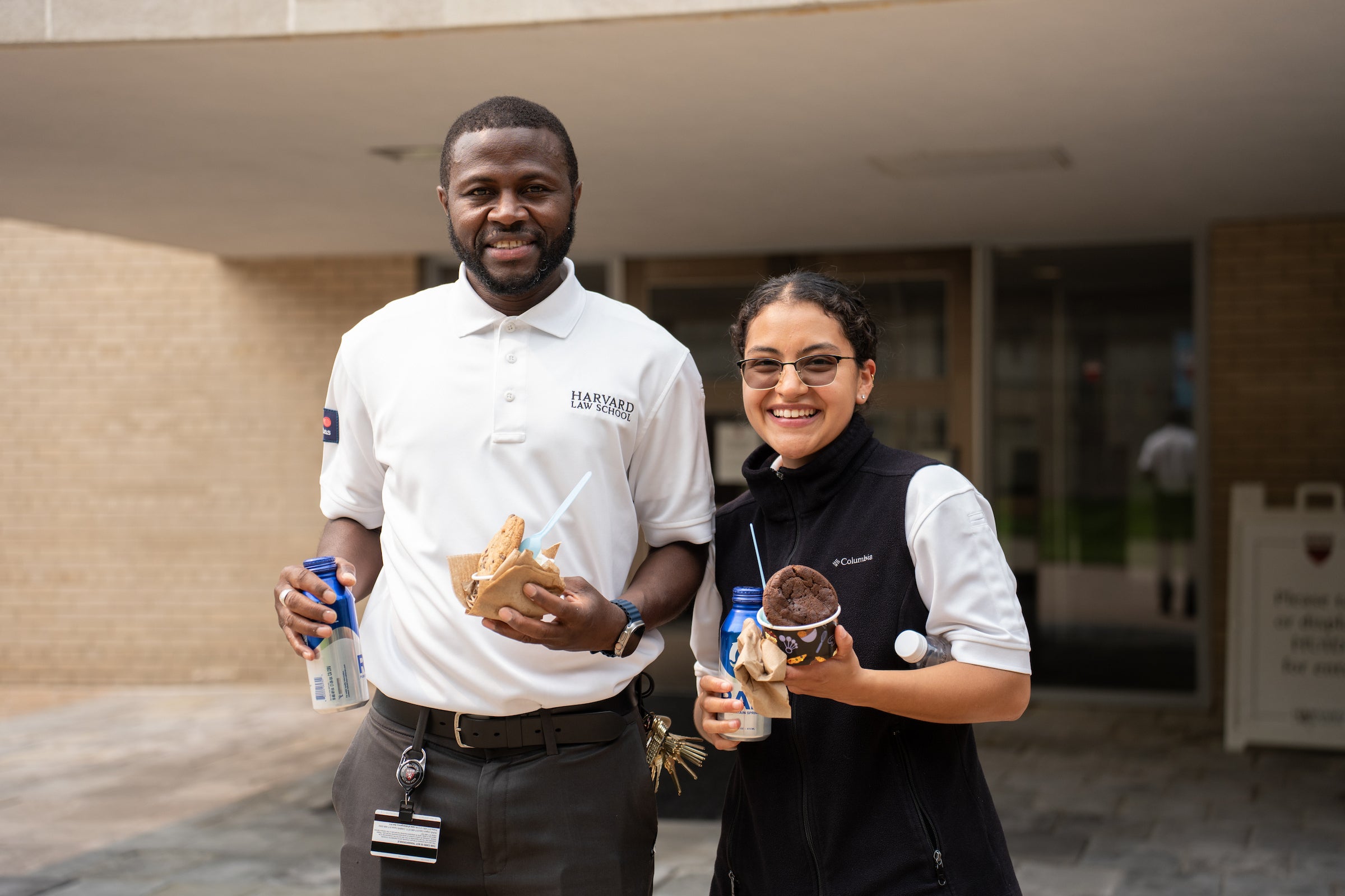 Two individuals in Harvard Law School attire stand outdoors holding food and drinks, smiling for the camera.