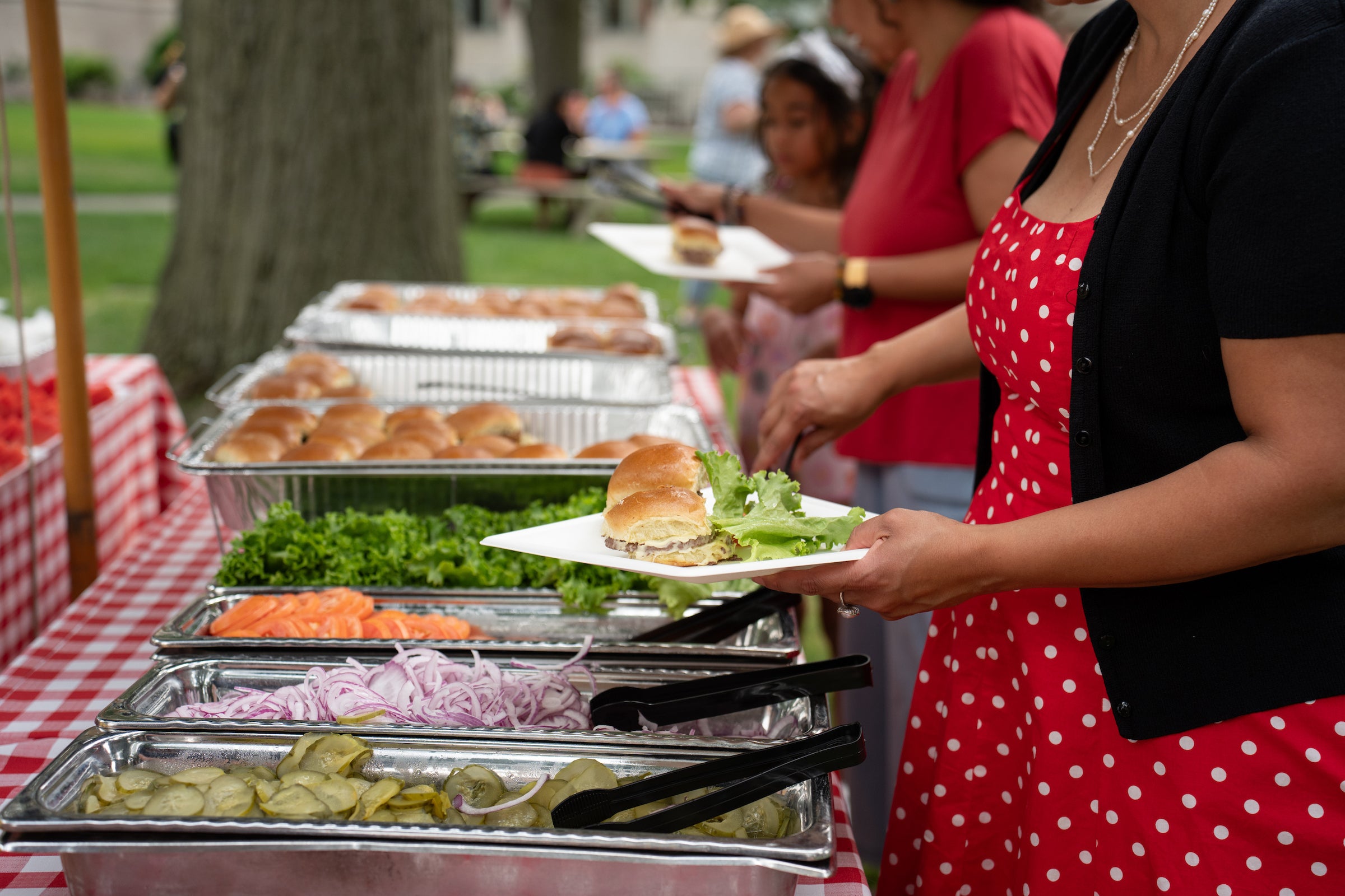 A person in a red polka-dot dress selects a sandwich from a buffet table laden with fresh toppings and bread rolls.