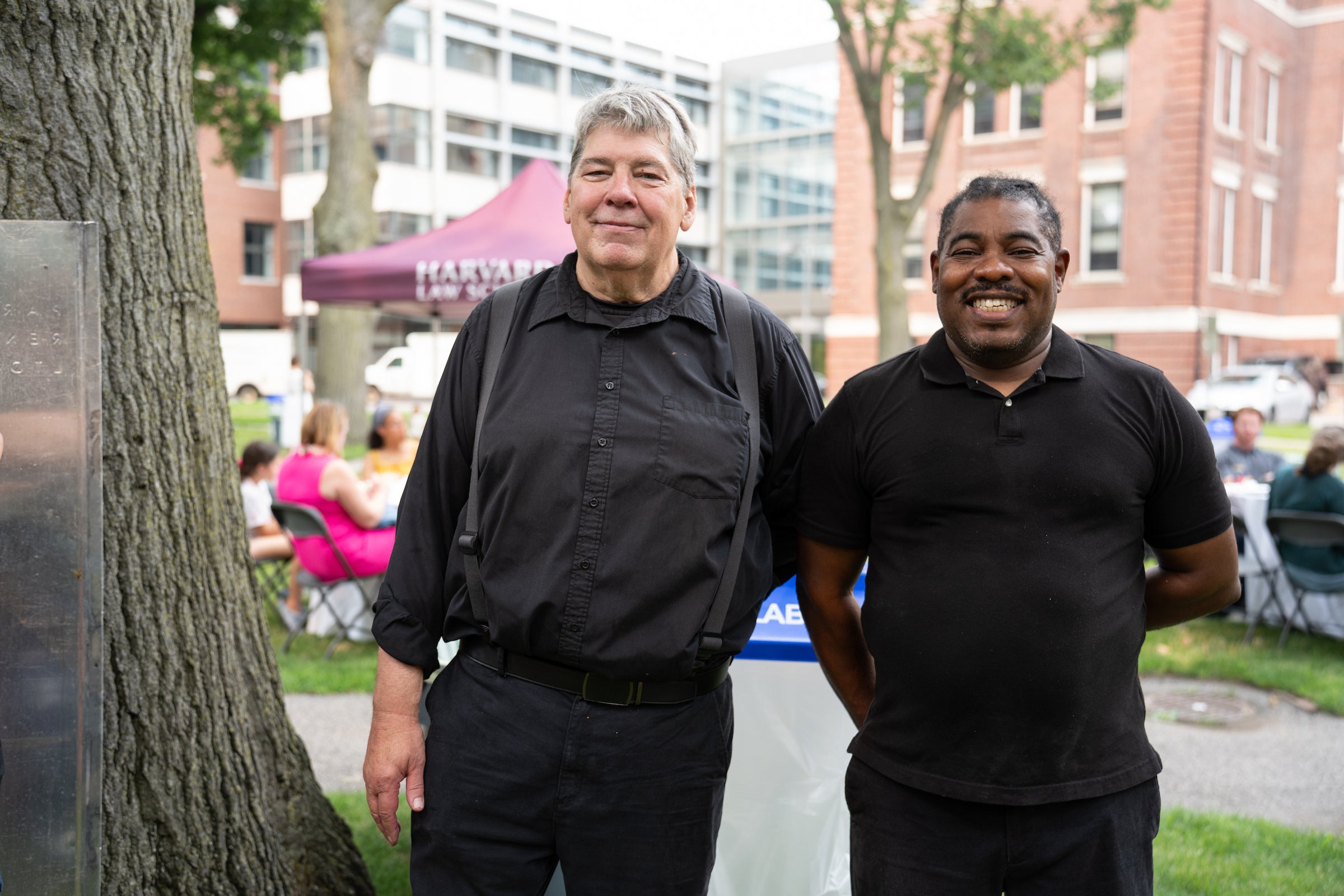 Two individuals standing outdoors smile for the camera, with a Harvard Law School tent and campus buildings visible in the background.