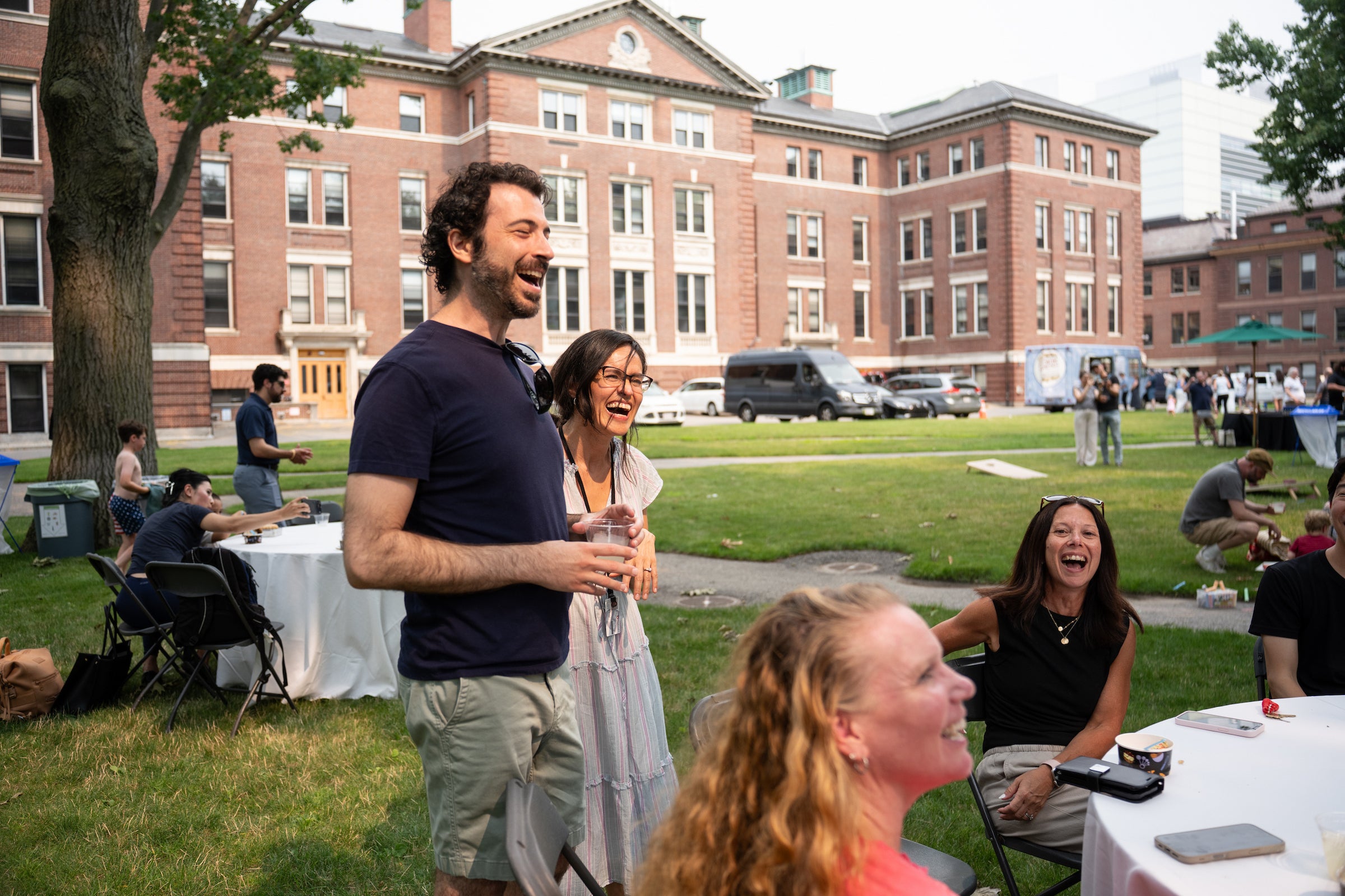A group of people laugh and socialize outdoors on a grassy lawn in front of a large brick building.