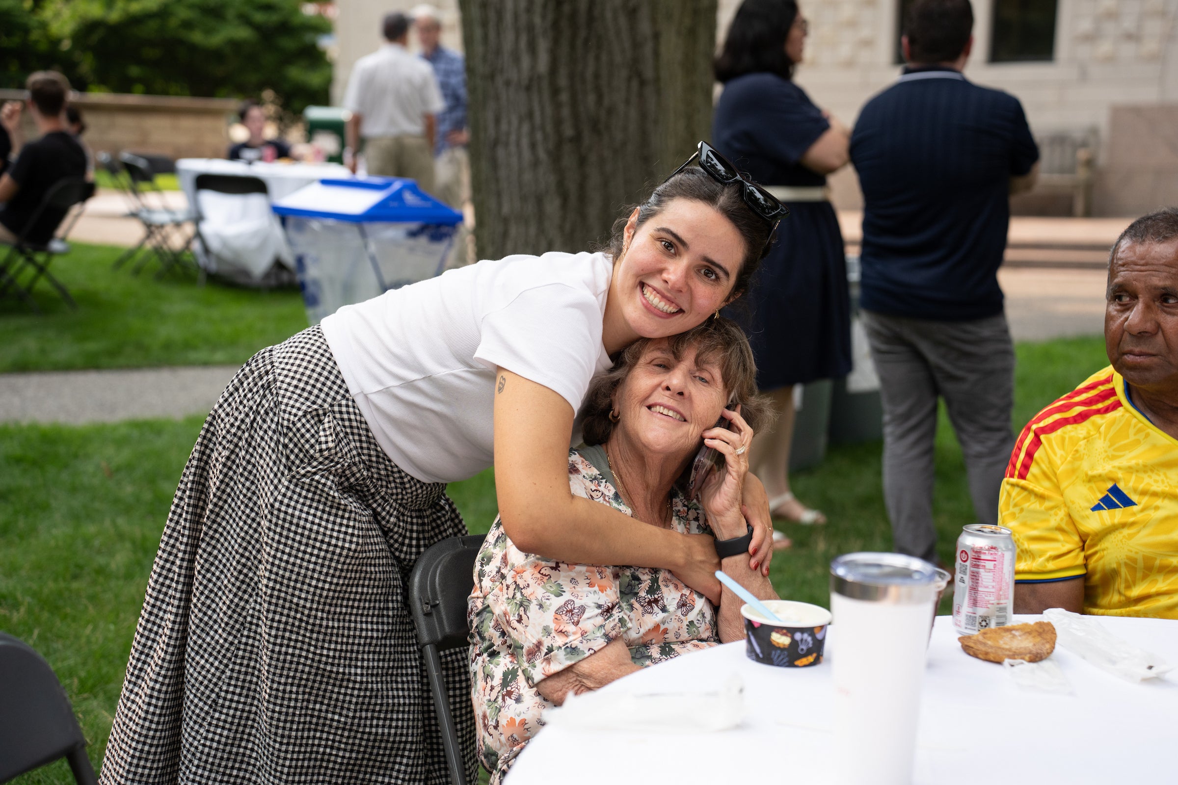 A standing person leans over a seated individual in a hug at an outdoor table, both smiling, while others converse in the background.