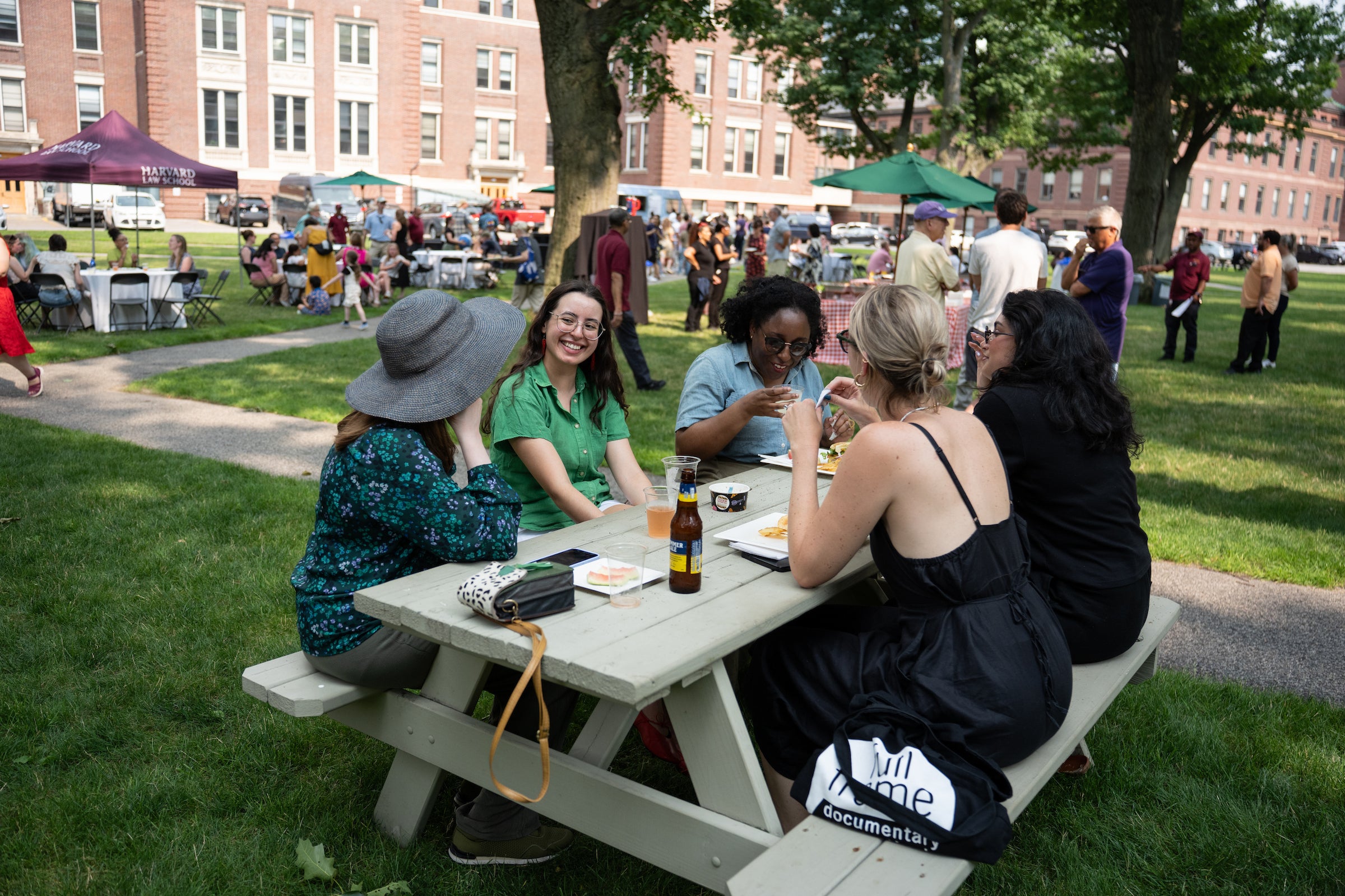 A group of individuals enjoys a meal together at a picnic table in a grassy outdoor area, surrounded by trees and other people at an event.