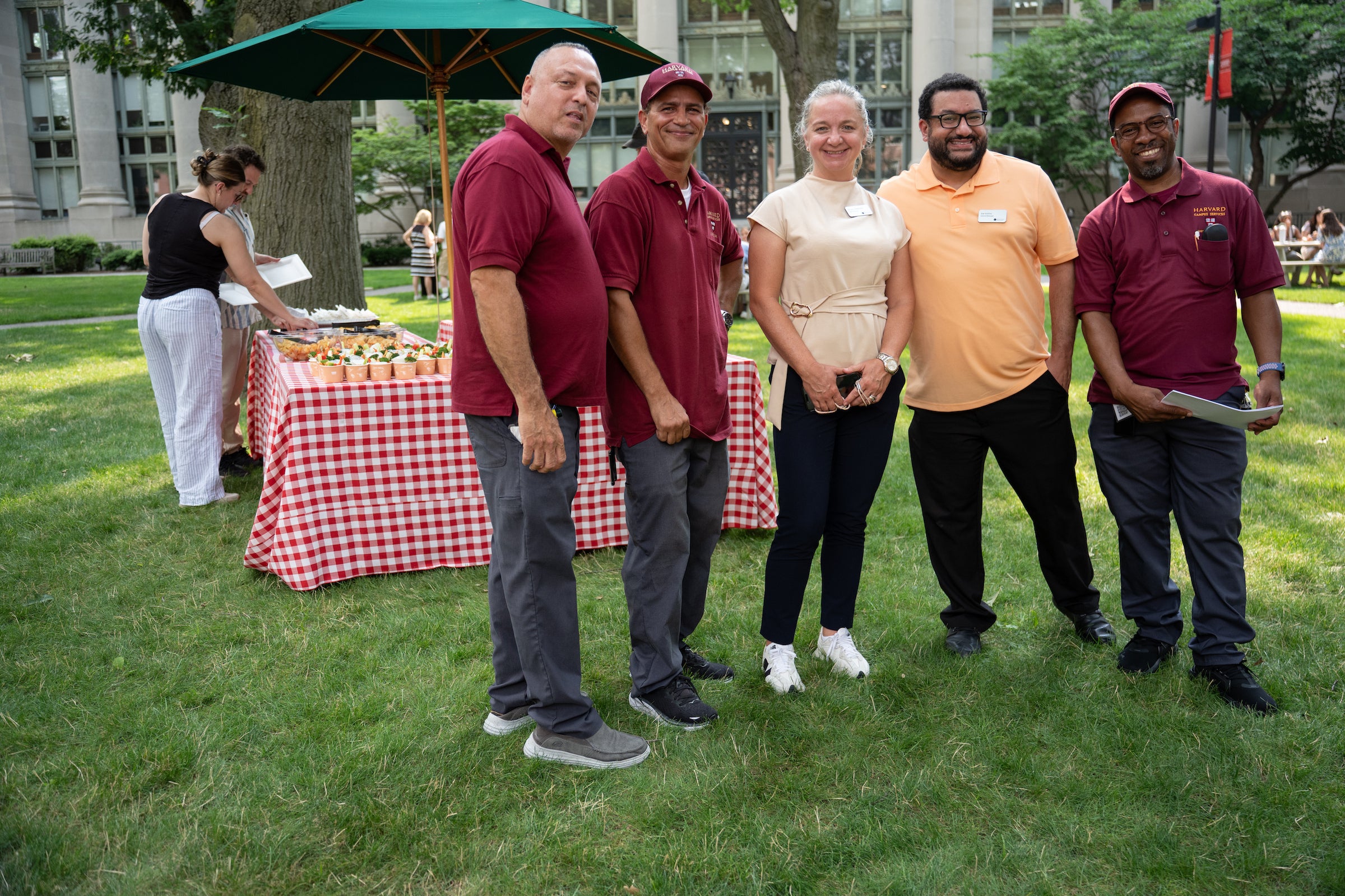 Five individuals stand together on a grassy lawn, with a red-and-white checkered table holding food behind them.