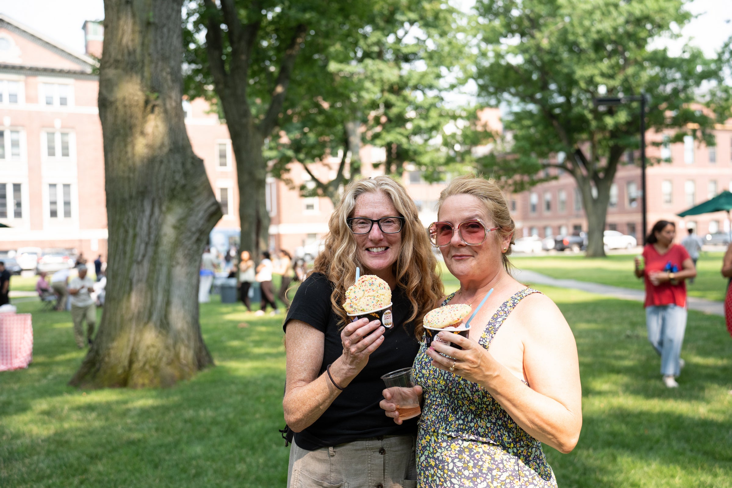 Two individuals smile while holding ice cream cups with trees and buildings in the background.