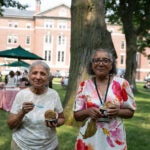 Two individuals enjoy ice cream outdoors in a grassy area with a brick building and trees in the background.