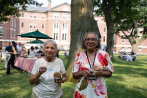 Two individuals enjoy ice cream outdoors in a grassy area with a brick building and trees in the background.
