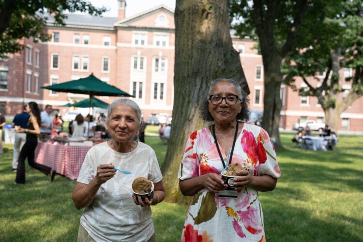 Two individuals enjoy ice cream outdoors in a grassy area with a brick building and trees in the background.