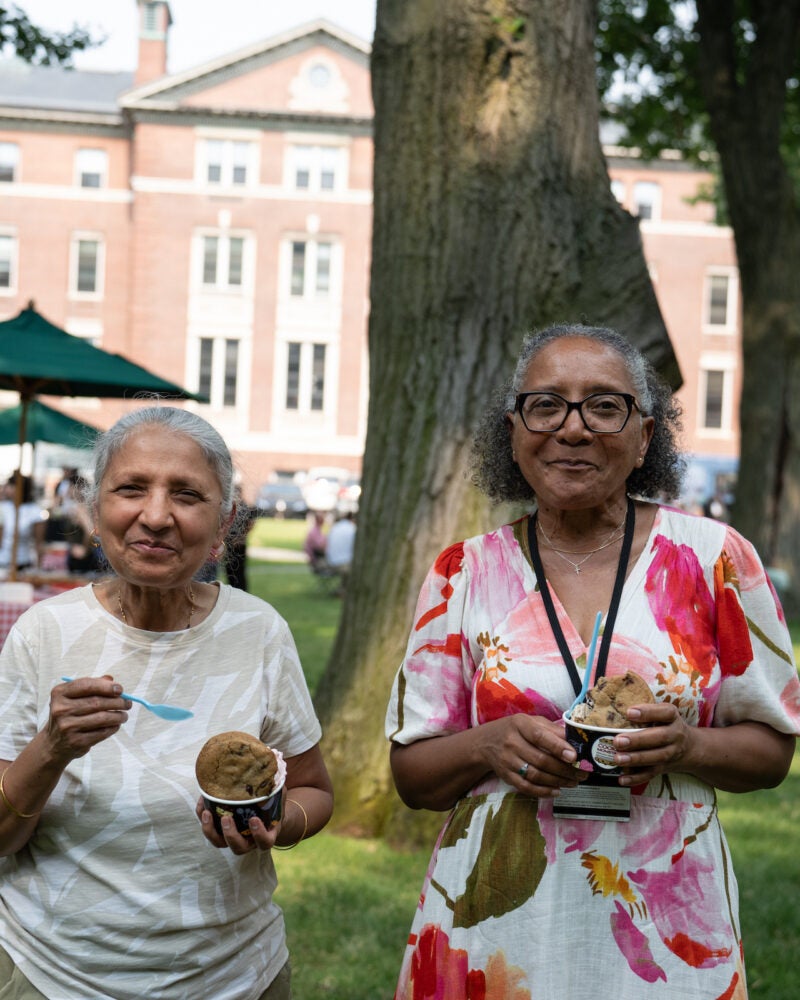 Two individuals enjoy ice cream outdoors in a grassy area with a brick building and trees in the background.