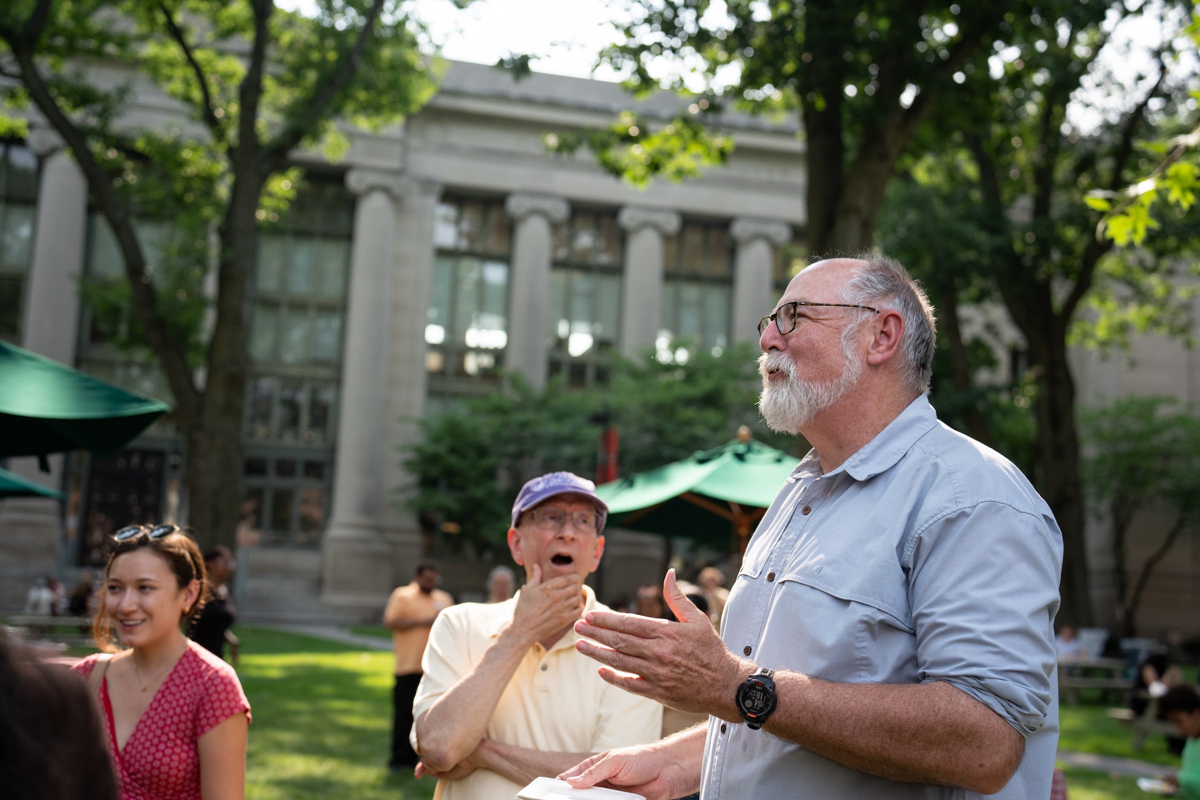 An individual with a white beard and glasses gestures while speaking to others in an outdoor setting, with a classical building and trees in the background.