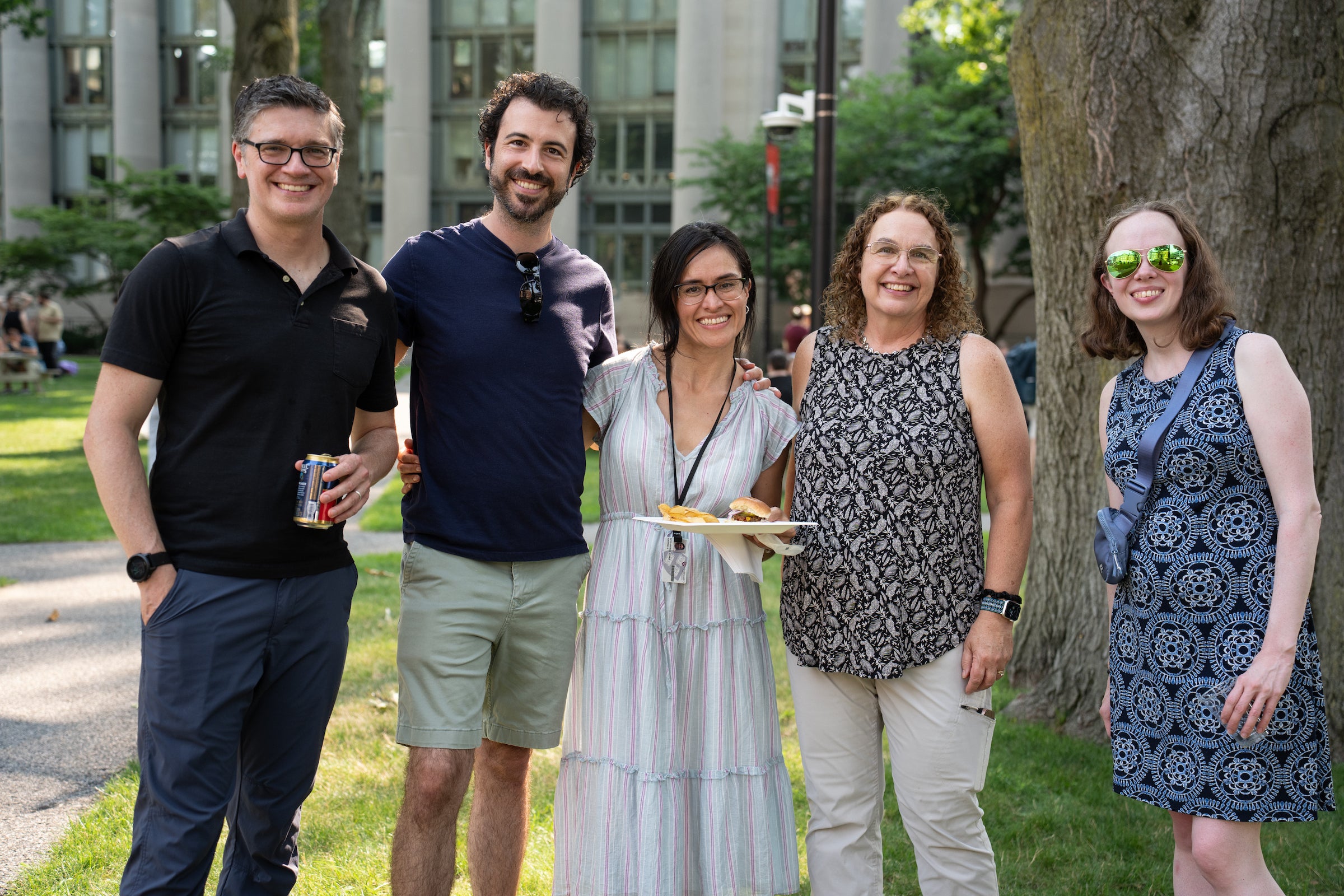 Five people smile for a photo outdoors, with one holding a plate of food and another holding a beverage.