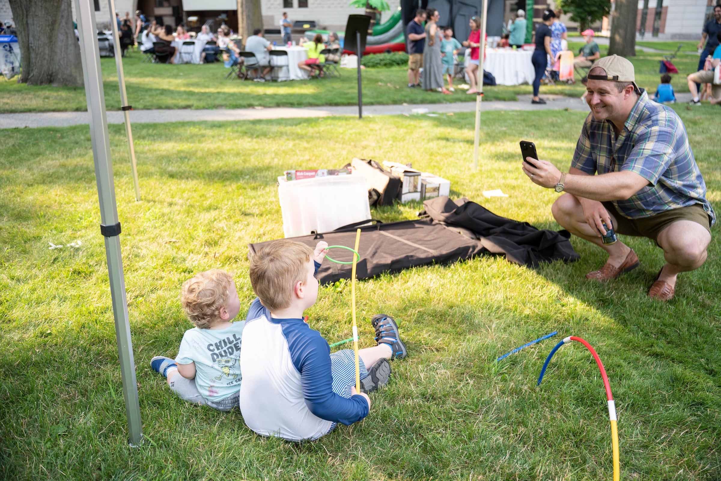 A person crouches on grass taking a photo of two children playing with colorful hoops near a tent.