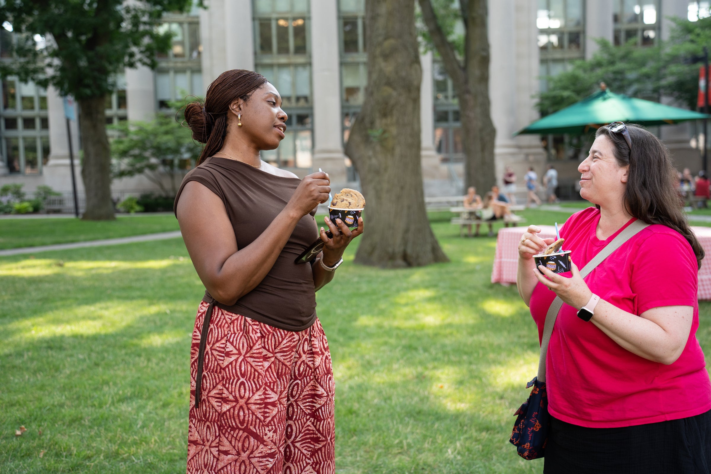 Two individuals converse while eating ice cream, with trees and a building with columns in the background.
